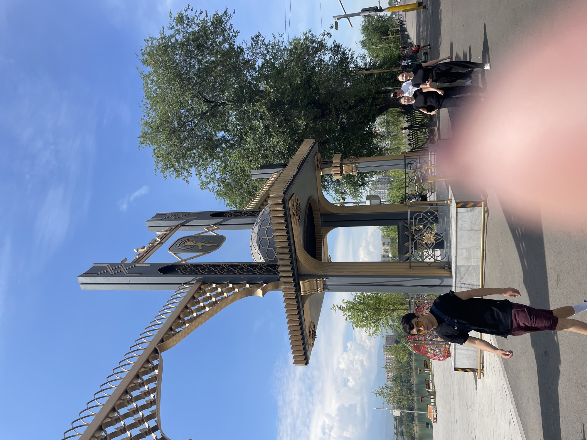 This is a street-level, medium shot image of a decorative archway structure with a man and a group of women walking on a paved walkway. The archway is ornate, with gold and dark gray elements and decorative patterns. A large, leafy tree is prominent behind the archway. In the background, there is a body of water, buildings, and mountains under a clear blue sky with some white clouds. The man in the foreground is wearing a black t-shirt, maroon shorts, sunglasses, and white socks. He is walking from left to right. The group of women are walking on the walkway to the right of the man, all wearing black outfits. The scene appears to be a public park or promenade, likely in the city of Almaty, Kazakhstan, given the presence of the decorative archway which is characteristic of the city's modern architecture and public spaces. The foreground is blurred by a pinkish object, possibly a finger.