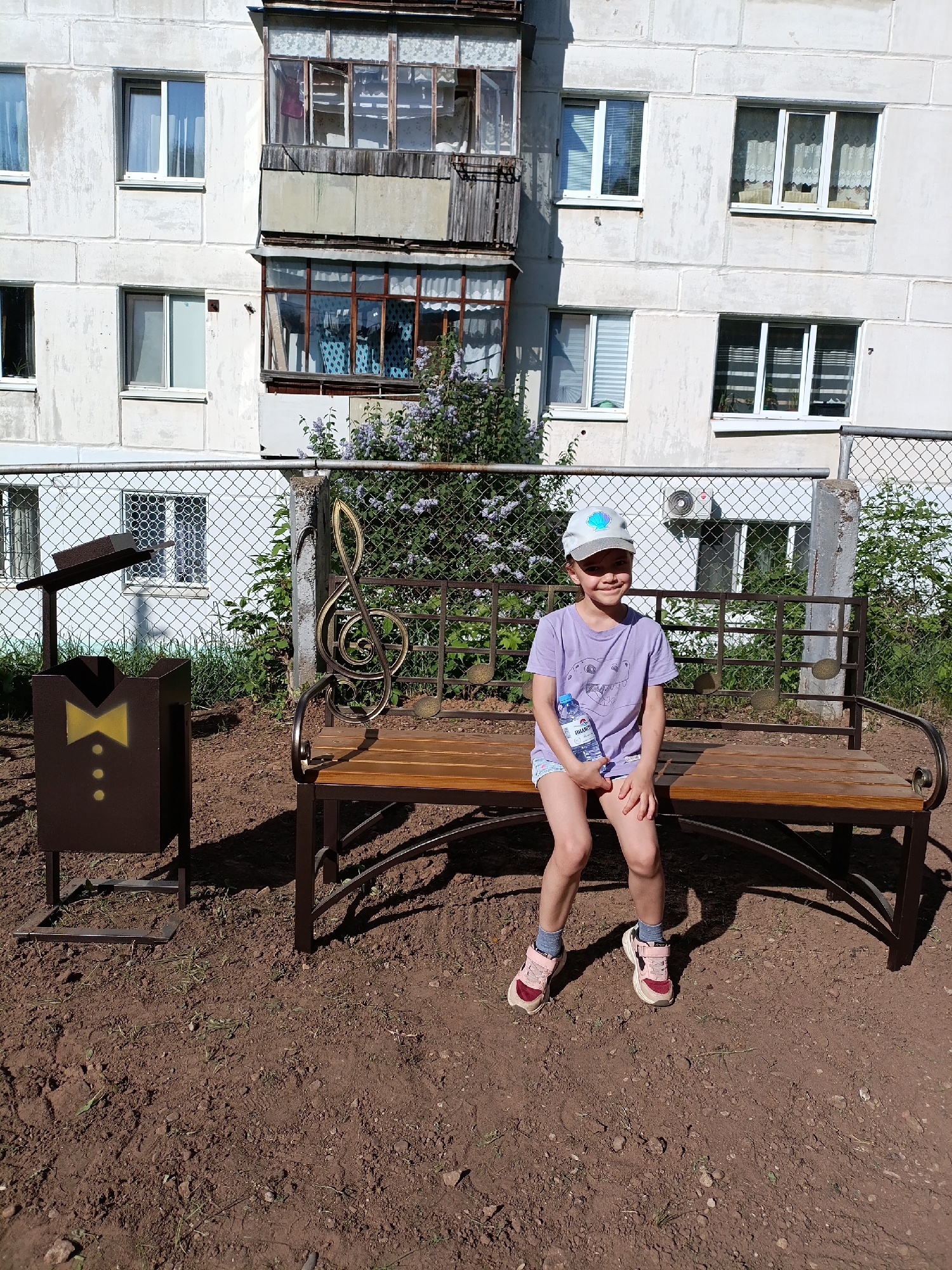 A girl is sitting on a park bench in Chaikovsky, Russia. The bench has a musical treble clef design on the backrest. To the left of the bench is a brown trash can resembling a tuxedo. Behind the bench is a chain-link fence and a white apartment building with multiple windows and an enclosed balcony. The ground is brown dirt. The girl is wearing a purple t-shirt, shorts, a cap, and pink sneakers. She is holding a water bottle. The scene is well-lit, suggesting it's daytime.