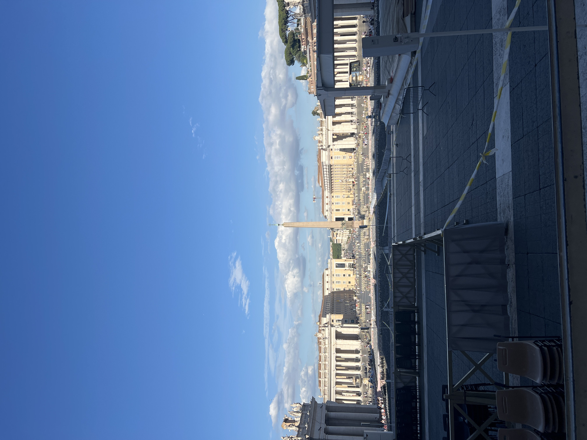 The image depicts an expansive view of St. Peter's Square in Città del Vaticano, Italy, under a clear blue sky with scattered white cumulus clouds. The foreground consists of dark gray paving stones, delineated by various temporary structures. On the immediate left, several stacks of light brown plastic chairs are visible. Moving further into the square, dark-colored chairs are arranged in rows within designated, fenced-off sections, indicating event seating. Yellow and white striped caution tape is present on the lower right, strung between poles.
In the midground, the iconic St. Peter's Square stretches out, featuring the prominent Vatican Obelisk centrally positioned, topped with a cross. To the left and right, parts of the semicircular colonnades, adorned with statues along their rooftops, frame the square. Beyond the left colonnade, a portion of St. Peter's Basilica is visible. Numerous people are distributed across the square, some walking, others seated in the prepared sections. Various cream-colored historic buildings, characteristic of the Vatican, line the perimeter beyond the colonnades. A fountain is visible on the left side of the square.
The background shows additional buildings and distant features, with distinctive umbrella pine trees visible on the far right. The scene is illuminated by bright daylight, casting clear shadows. The overall impression is of an iconic public space prepared for a large gathering or event.
