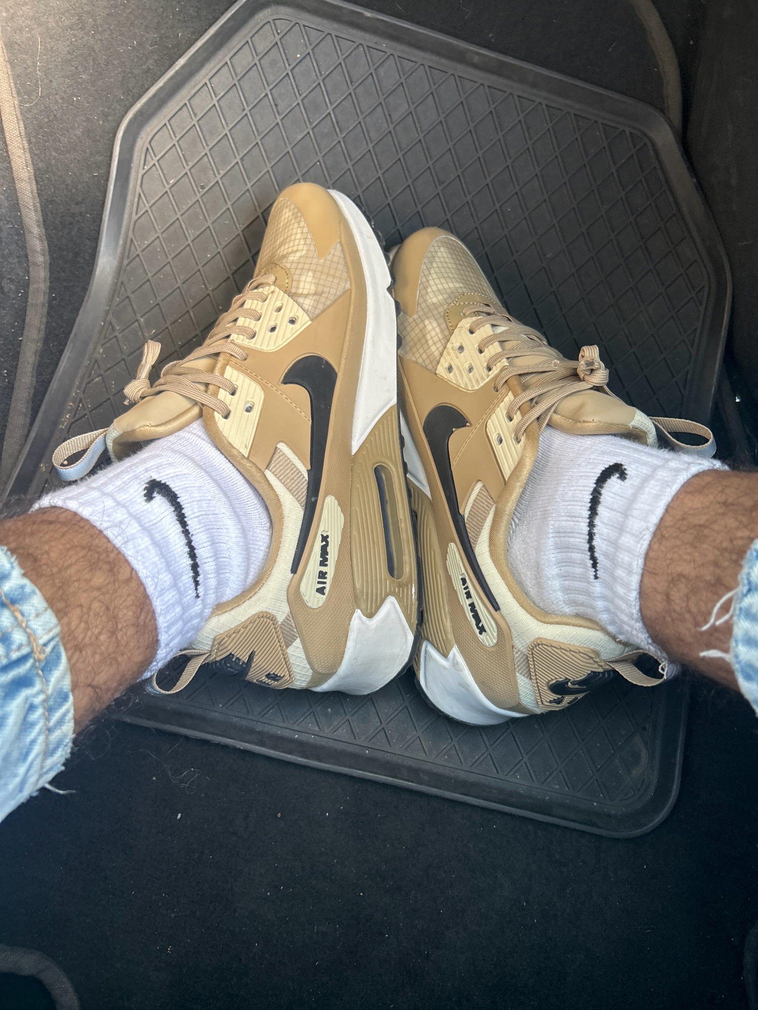 The image shows a close-up, overhead view of a person's feet wearing tan and white Nike Air Max sneakers and white crew socks with black Nike swooshes. The shoes are resting on a black rubber floor mat in what appears to be the interior of a vehicle, possibly a car, given the texture of the surrounding dark fabric. The sneakers feature tan uppers with a checkered pattern on the side panels, white soles, and black Nike swooshes. The socks are plain white with a black Nike swoosh logo visible on each. The lower legs and upper thighs of the person are partially visible, clad in light-wash denim shorts. The overall setting suggests a casual, personal moment captured inside a car, possibly parked. The image does not provide any specific clues or context about the city of Montebello della Battaglia, Italy.