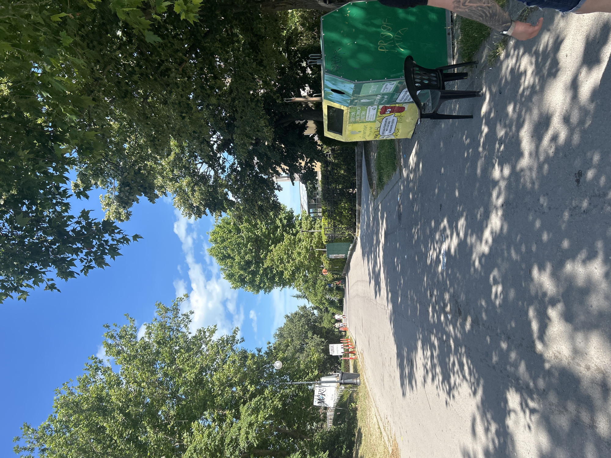 This media file depicts a sunny day in Varna, Bulgaria, likely during the warmer months given the lush green trees and clear blue sky. The scene is an outdoor public space, possibly a park or a street lined with trees.
The primary focus is on a large, multi-compartment recycling bin, predominantly green and yellow, with various labels and graffiti. Adjacent to the bin is a black plastic chair, suggesting a resting spot or a temporary placement. In the background, a paved pathway or road stretches into the distance, shaded by the dense foliage of mature trees. The shadows of the leaves cast intricate patterns on the asphalt.
Further down the path, several orange traffic cones are visible, hinting at potential roadwork or a delimited area. A sign for "CLASSIC HOTEL" can be spotted among the trees, providing a geographical clue. Another sign reads "MORE 50 м," likely indicating a distance to a point of interest.
A person's arm with intricate tattoos is visible on the right side of the frame, reaching towards the recycling bin, suggesting an interaction with the environment. The overall mood is relaxed and casual, bathed in the bright sunlight. The visible text on the recycling bin includes "PROOF," "Стъкло" (Glass), "МЕТАЛ" (Metal), and some Cyrillic phrases in smaller print, including what appears to be instructions or slogans related to recycling. The presence of the bin and the signs indicate a developed urban or semi-urban area.