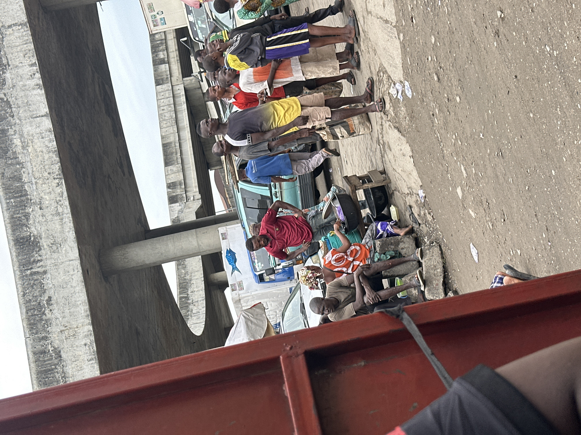 This media file captures an active outdoor scene in Agani, Nigeria, under an overcast daytime sky. A large concrete overpass or bridge spans the left side of the frame, providing a substantial architectural element.
Numerous people, mostly adult males, populate the scene. A prominent group of about ten men stands in a line towards the center-left, dressed casually in t-shirts, shorts, and trousers, appearing to be waiting or gathered. Near this group, a sack is visible with the text "POUDR".
In the lower parts of the frame, other individuals are resting or sitting. A man in a red shirt is seen near a light blue truck, which features a distinctive blue fish logo and the partially visible text "LEVEM" on its side. To the right, another man in a light brown shirt and a woman in an orange and white patterned dress are seated on the ground or low structures, amidst plastic buckets, discarded papers, and other miscellaneous items.
The ground surface is a mix of concrete and dirt, showing signs of wear. A red metal structure occupies the immediate foreground, suggesting the image was captured from inside a vehicle or behind a barrier. The overall impression is a snapshot of daily life in a bustling market or roadside area, with no strong emotions discernible among the calm and indifferent individuals.