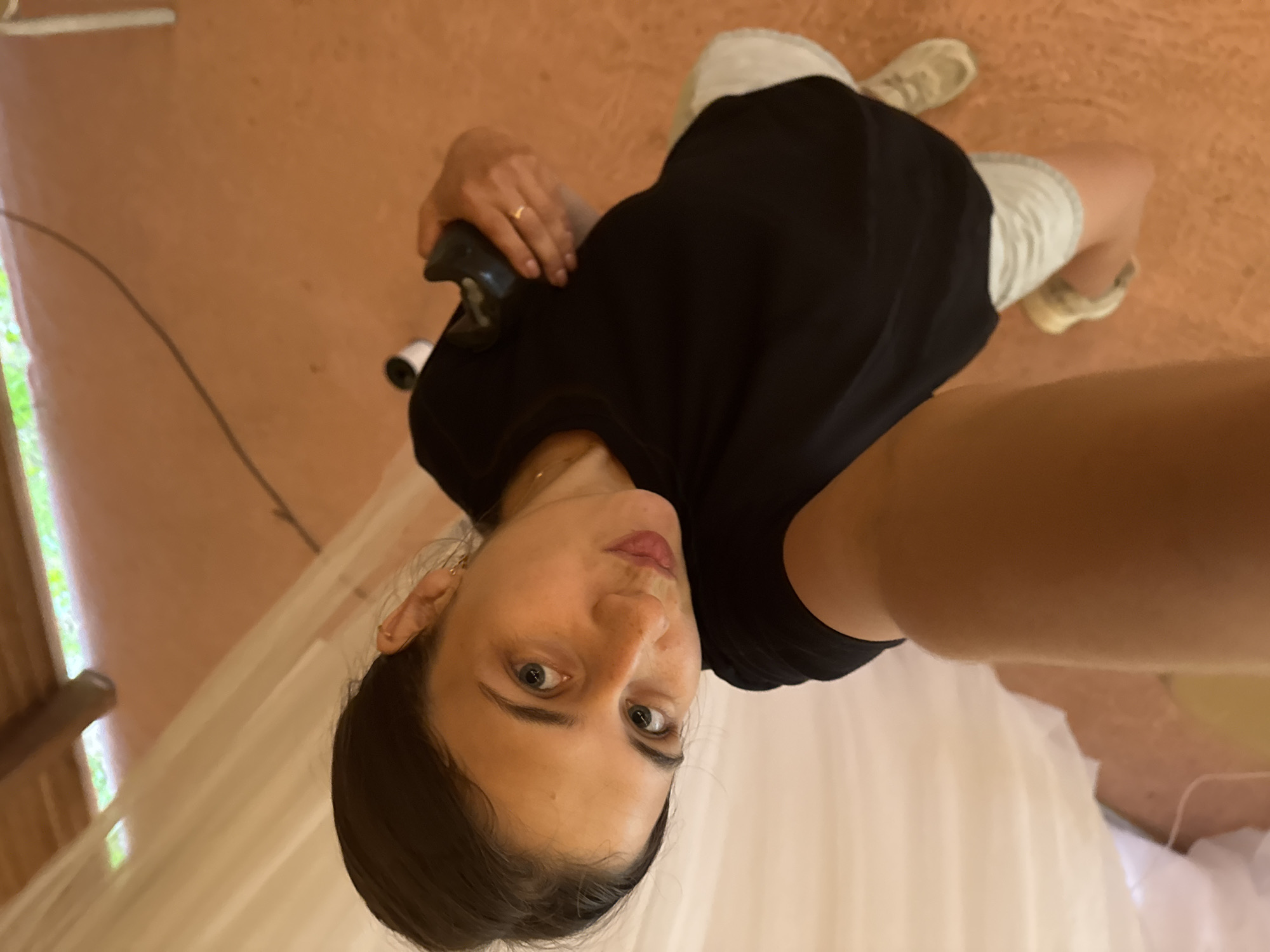 An upward-angle, full-body, selfie-style photograph captures a woman in a black t-shirt and light-colored shorts. She has dark hair pulled back, and is wearing light-colored sneakers. She is holding a black object in her right hand, and a thin gold ring is visible on her ring finger. The environment appears to be an indoor space with a tiled or concrete floor. A white sheer fabric is visible to the left of the frame, and a wooden beam is present at the top of the image. The lighting suggests it is daytime, and the overall impression is of a casual, personal moment.