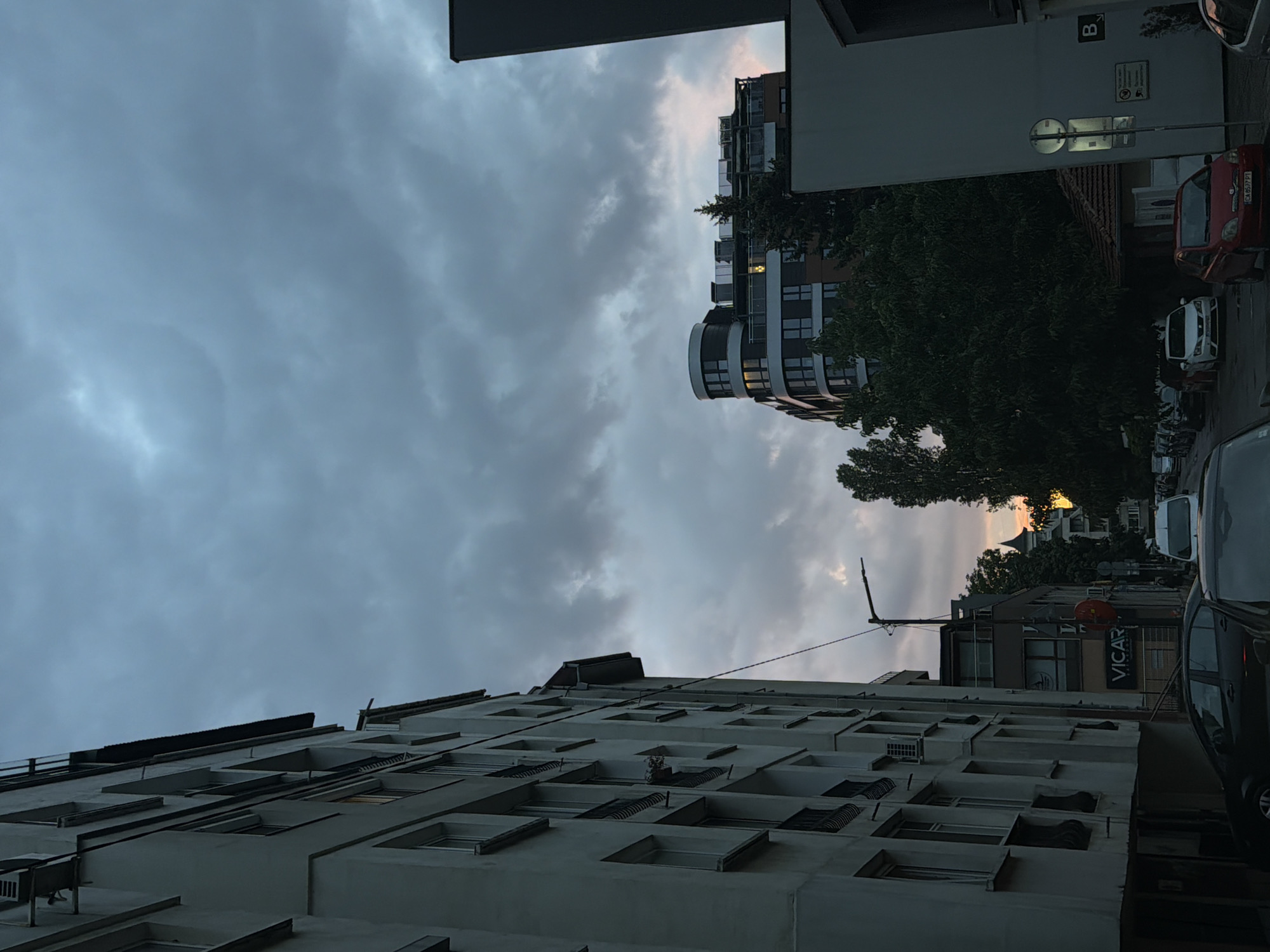 The image shows a street scene in Sofia, Bulgaria, at dusk. A tall, light-colored apartment building occupies the left side of the frame, its facade marked by numerous windows and balconies. The sky above is filled with heavy, gray clouds, with a faint hint of pinkish light peeking through on the right. In the mid-ground, a modern, curved building with dark-tinted windows stands out, partially obscured by lush green trees. Further down the street, smaller buildings and parked cars are visible, including a red hatchback and a white hatchback. A street lamp is positioned mid-frame, and traffic signs are present. The overall atmosphere is somewhat somber due to the overcast sky and the late hour.