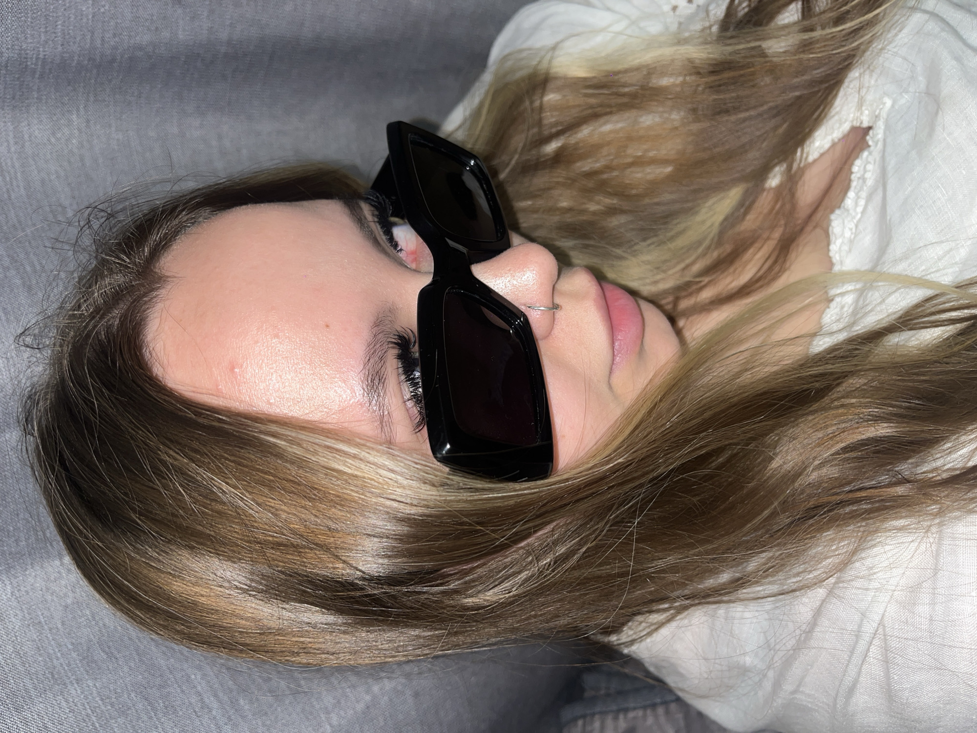 A woman lies supine on a gray upholstered surface. She wears black rectangular sunglasses and has a silver nose ring. Her hair is light brown with blonde highlights. Part of a white, lace-trimmed garment is visible near her head. The location in Krasnodar, Russia is not determinable from the image.