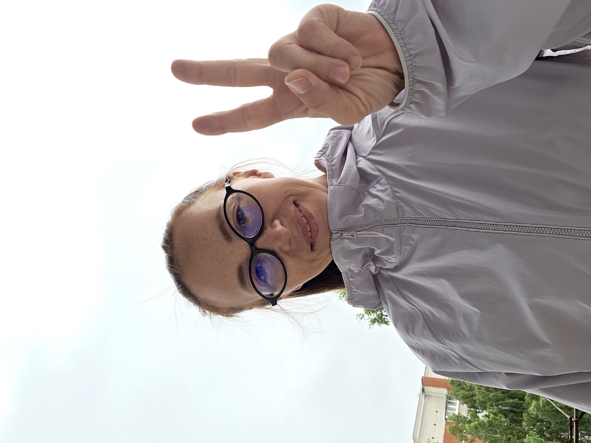 This media file is a selfie taken outdoors with a light purple jacket. The subject is a person wearing round-framed glasses and a light purple jacket, making a peace sign with their right hand. The background is a bright, overcast sky, suggesting cloudy weather. There are some green trees and a building visible in the lower right portion of the frame, indicating an urban or suburban setting. The photo is taken from a low angle, looking up at the subject. The time of day is not clearly identifiable, but the lighting suggests it is daytime. No text is visible in the image. The overall mood appears cheerful and casual.