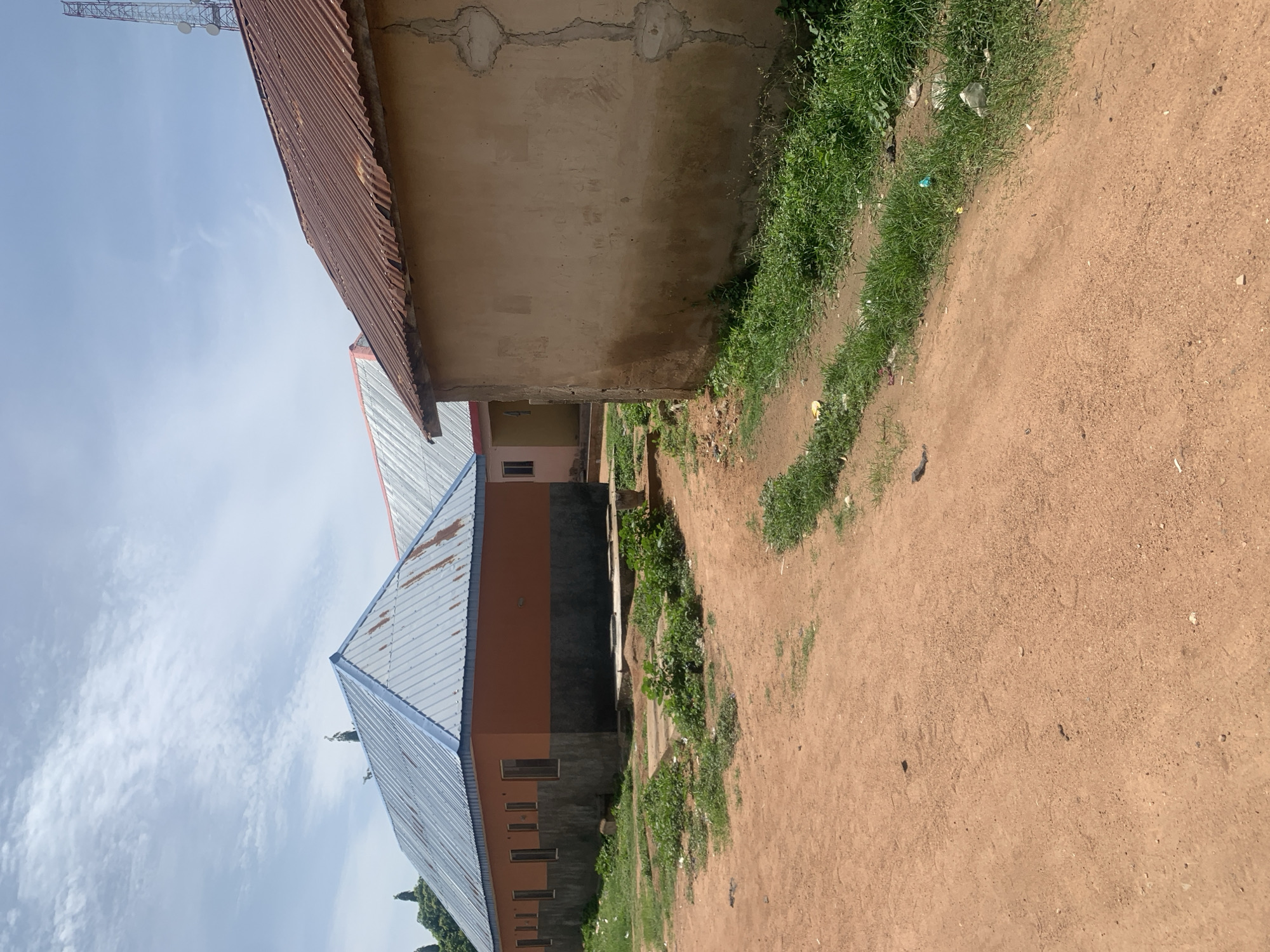 A scene unfolds in the city of Jauro Yinu, Nigeria, under a sky adorned with wispy clouds. In the foreground, a dirt road, tinged with the warm hues of dried earth, curves gently, its surface marked by faint tire tracks. To the right, a dense patch of green grass borders the road, punctuated by scattered pieces of litter.
Dominating the right side of the image is a building with a weathered, tan-colored facade, its rough texture hinting at age and exposure to the elements. A prominent vertical crack runs down its surface, adding to its rustic appearance. To the left, a building with an orange-colored upper story and a dark gray base stands prominently. Its corrugated metal roof, painted a light blue, gleams in the sunlight, showing some signs of rust. Several rectangular windows are visible along its orange wall, framed by dark gray structures.
Behind these structures, another building with a red corrugated metal roof peeks out, its presence hinting at a larger complex. In the distance, a tall, red-and-white telecommunications tower with multiple platforms and antennas pierces the sky, a modern sentinel against the horizon. Scattered vegetation, including trees with dark green foliage, can be seen in the background, adding a touch of natural contrast to the man-made structures.