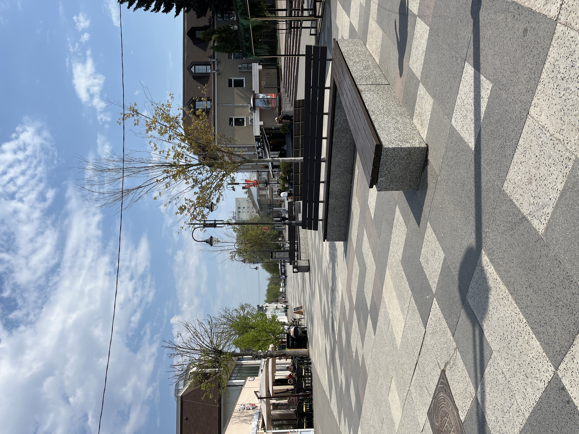 The image depicts a pedestrian walkway in Noginsk, Russia. The walkway is paved with alternating grey and white geometric patterned tiles. A series of benches with dark wooden slats and speckled stone supports line the walkway. Trees with sparse foliage are planted along the street. Buildings of varying heights and architectural styles border the walkway. Street lamps are visible. The sky is blue with scattered clouds. Some people are walking in the distance. The photograph is taken at an angle, with the walkway stretching into the distance. A manhole cover is visible in the foreground. A power line is visible against the sky.