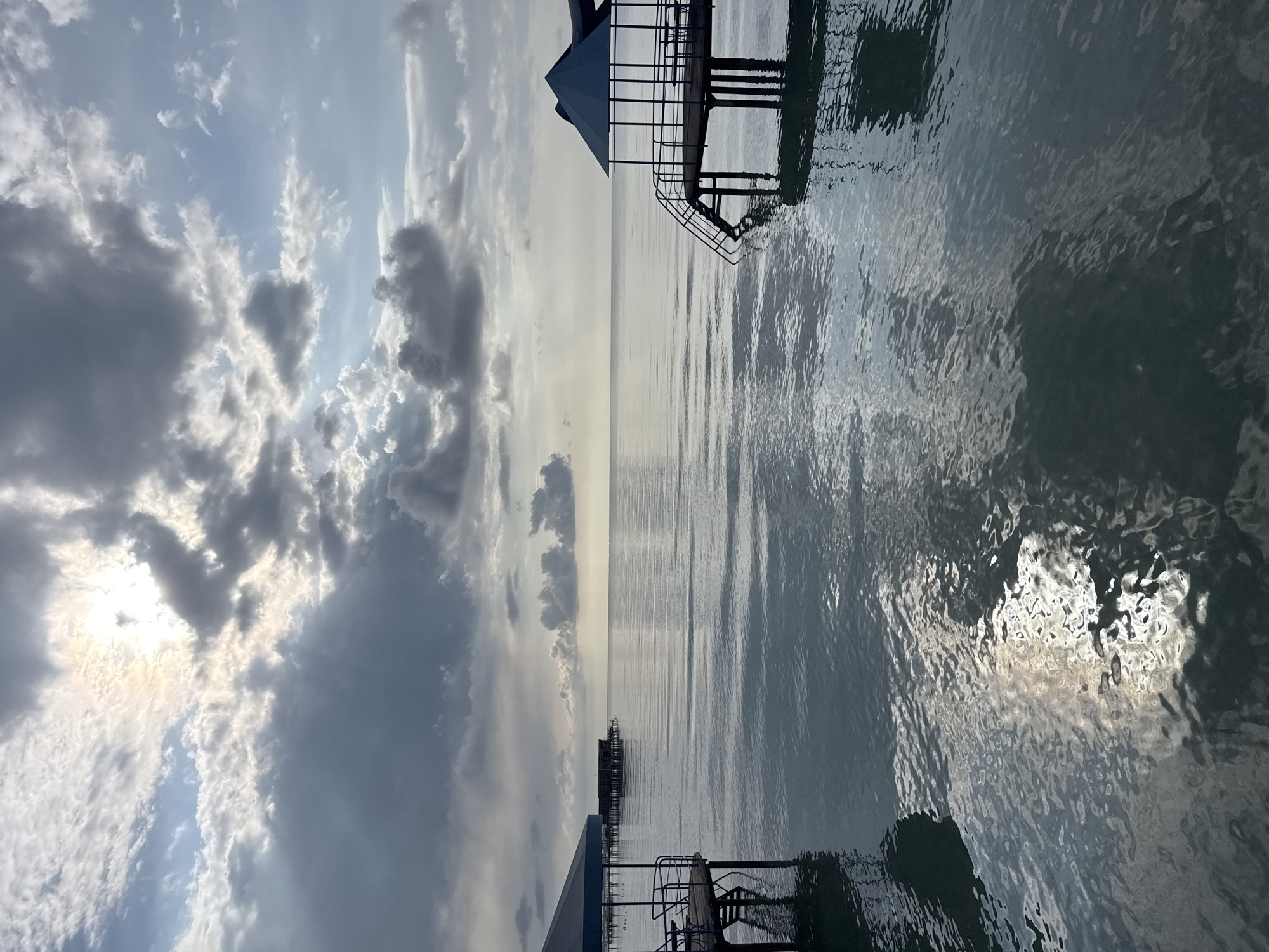 This is an outdoor, eye-level, medium shot of a lake scene at Sary-Oy, Kyrgyzstan. In the foreground, on the left, a wooden pier extends into the water. It is covered by a dark blue, angled canopy supported by thin metal posts. A railing of the same material encloses the pier, with two chairs visible on the decking. On the right, another pier with a similar dark blue, pyramidal canopy juts out over the water. This pier also has a railing and a set of stairs descending into the lake. The water is calm, with ripples reflecting the cloudy sky and the structures. In the background, across the lake, a row of dark, multi-story buildings on stilts are visible. The sky is filled with dramatic, dark clouds interspersed with patches of bright sunlight breaking through. The overall mood is serene and slightly overcast.