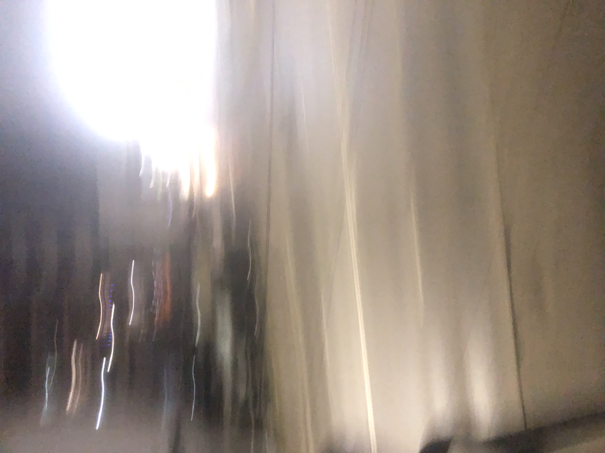 The image presents an abstract and dynamic urban scene, heavily affected by motion blur and strong lighting. On the left side, a powerful, overexposed light source dominates the upper quadrant, creating an intense glare that washes out details in that area. This bright light transitions into a softer, hazy luminosity extending towards the center of the frame.
To the right, the image features several distinct vertical lines and subtle tonal variations, suggesting the presence of a building facade, a wall, or window structures. The surfaces appear muted in color, possibly beige or light brown, and reflect some of the ambient light.
In the lower-left and central parts of the image, against a darker, indistinct background, a series of elongated, wavy vertical streaks of light are visible. These streaks are primarily white, with some faint blue and warm yellow-orange tones, strongly indicating artificial light sources such as streetlights or vehicle lights that have been stretched by camera movement.
No specific objects or individuals are clearly discernible due to the extensive blurring. The overall impression is that the photograph was taken from a moving vehicle or by a moving person, capturing the essence of an urban environment during night or twilight hours, with artificial illumination playing a significant role. There is no visible text in the image. The location context provided is Sofia, Bulgaria.