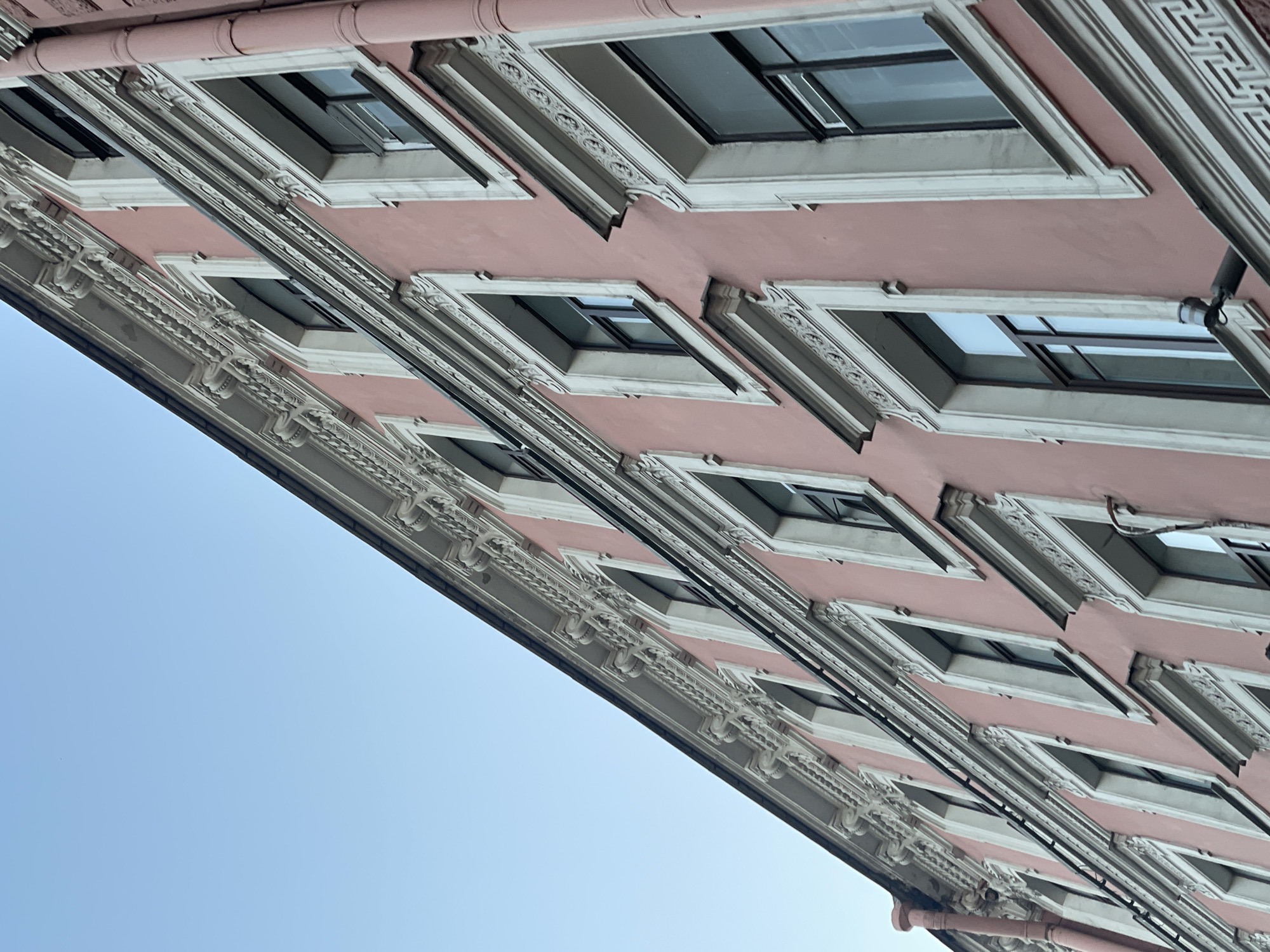 An upward shot reveals a pink building façade in Saint Petersburg, Russia. The structure exhibits an ornate, neoclassical style, identifiable by rows of rectangular windows bordered by white trim, embellished with intricate decorative moldings and keystones above each window. Grey linear architectural elements separate the windows. The building’s pink paint is contrasted by the clear blue sky occupying the lower left corner.