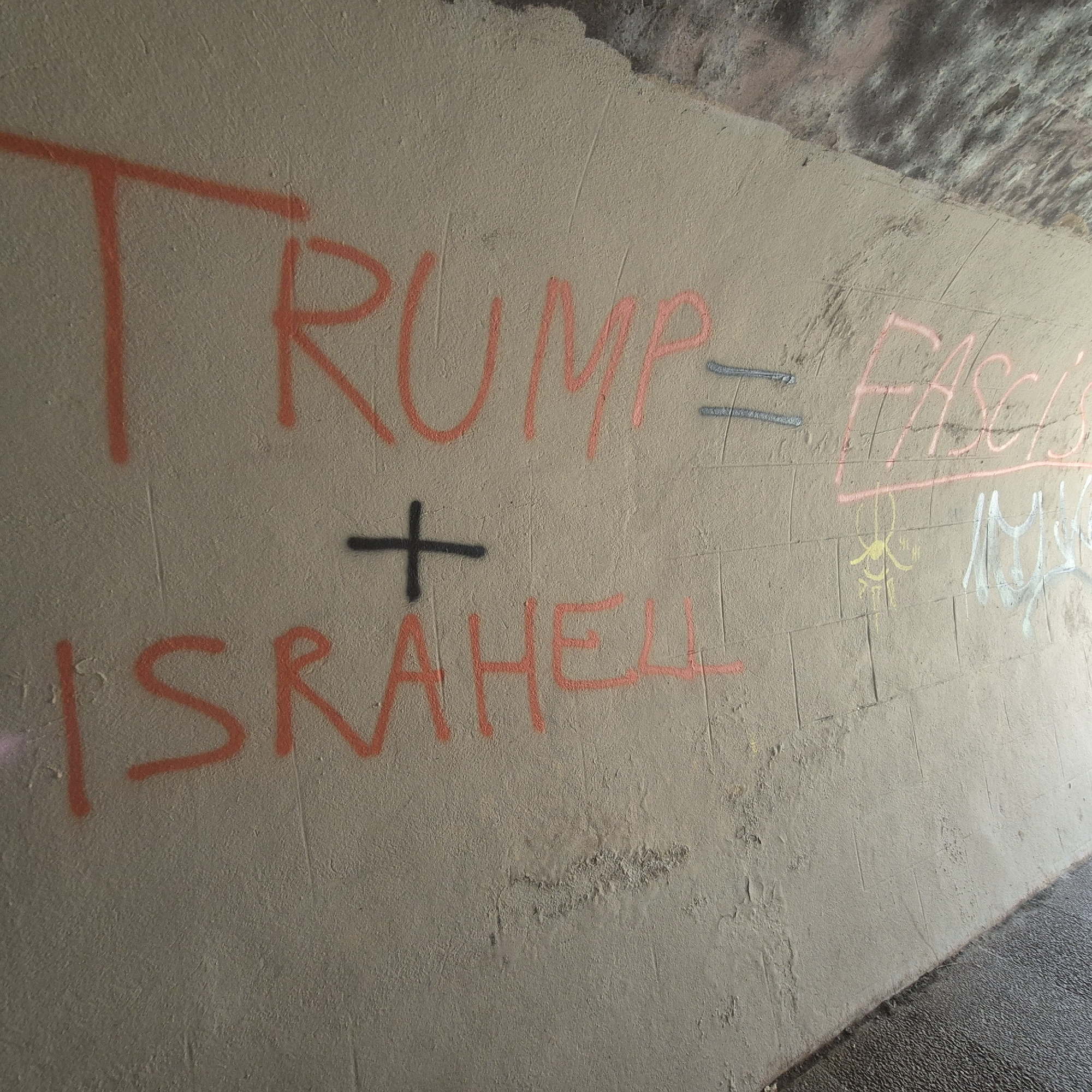Graffiti on a wall reads "Trump = Fascism + Israhell." The location is likely an underpass or tunnel in Strasbourg, France, given the architecture and graffiti style often found in urban environments. The graffiti appears to be spray-painted in red and other colors on a concrete wall. The wall also exhibits some pre-existing wear, including discolorations and damage. The floor is visible at the bottom right of the image.