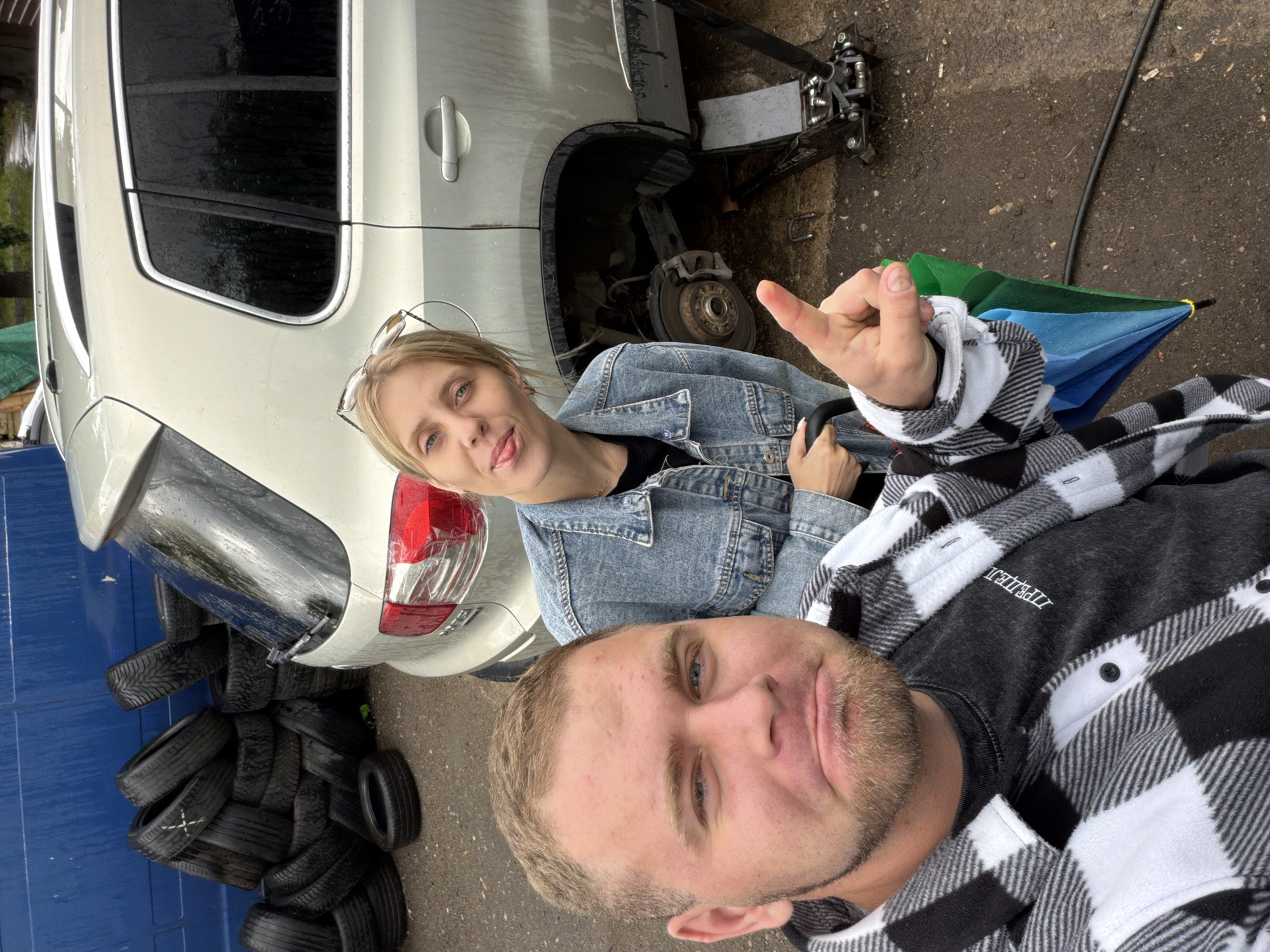 A man and a woman are taking a selfie next to a car that appears to be undergoing maintenance. The car is a silver SUV with its rear wheel removed, and a jack is visible underneath it. The woman is wearing a denim jacket and has blonde hair, and she is sticking her tongue out at the camera, with sunglasses on her head. The man is wearing a black and white checkered shirt and has a beard, and he is making a hand gesture with his fingers. A stack of tires is visible to the left of the car. A blue and green umbrella is also present in the frame. The ground is wet, suggesting it may have recently rained or is raining, and the overall lighting indicates it is daytime. The background includes a blue wall and some greenery, possibly indicating an outdoor setting like a garage or a yard. The people's expressions suggest a lighthearted and perhaps slightly mischievous mood, possibly related to the car repair. The word "ZIPEJJ" is visible on the man's shirt.