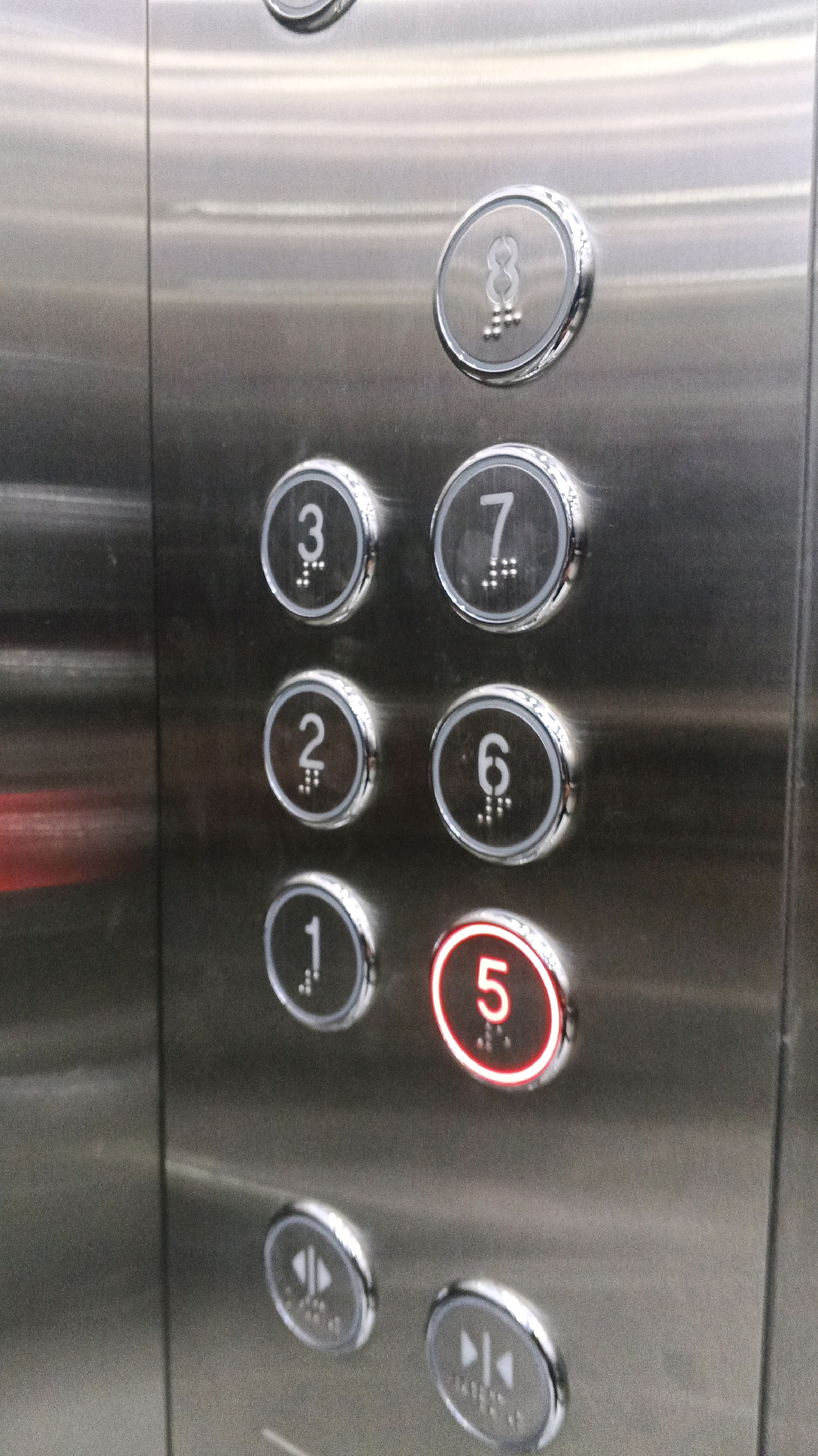 The image shows a close-up of elevator buttons on a stainless steel panel. The buttons are round and metallic, with numbers and Braille dots indicating their function. Several buttons are visible, including those for floors 8, 3, 7, 2, 6, 1, and 5. The button for floor 5 is illuminated with a red light, indicating it is currently selected or active. Below the numbered buttons are two smaller, circular buttons with symbols representing "open doors" and "close doors." The background is a brushed metal surface, characteristic of an elevator interior. The image does not provide any information about the city of Semarang, Indonesia, or any people or actions. It is a static shot of an elevator control panel.