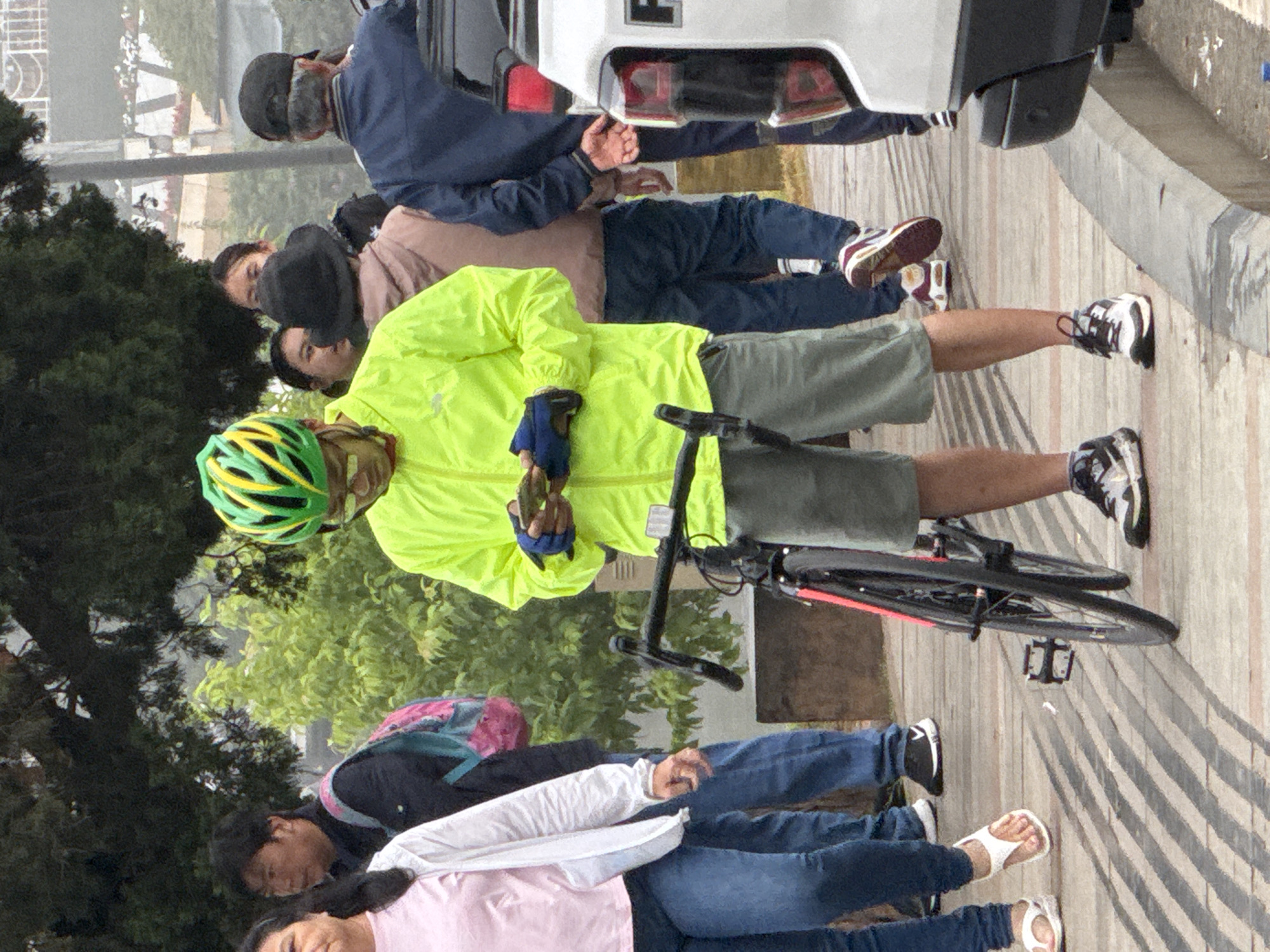 A group of people are walking along a paved pathway in Da Lat, Vietnam. A man in the foreground is wearing a bright yellow jacket, grey shorts, a green and yellow helmet, and black and white sneakers. He is holding a phone in his hands and looking down at it. A bicycle with a black frame and red accents is standing next to him, with its front wheel turned to the left.
To the left of the man, a woman in a pink shirt and white jacket is wearing blue jeans and white sandals. Behind her, a man in a dark jacket and a backpack with pink and blue straps walks past. Further behind them, several other individuals are visible, including a woman wearing a black hat and a man in a navy blue jacket. The background shows trees and some buildings. The pathway appears to be made of wooden planks with a rough texture.