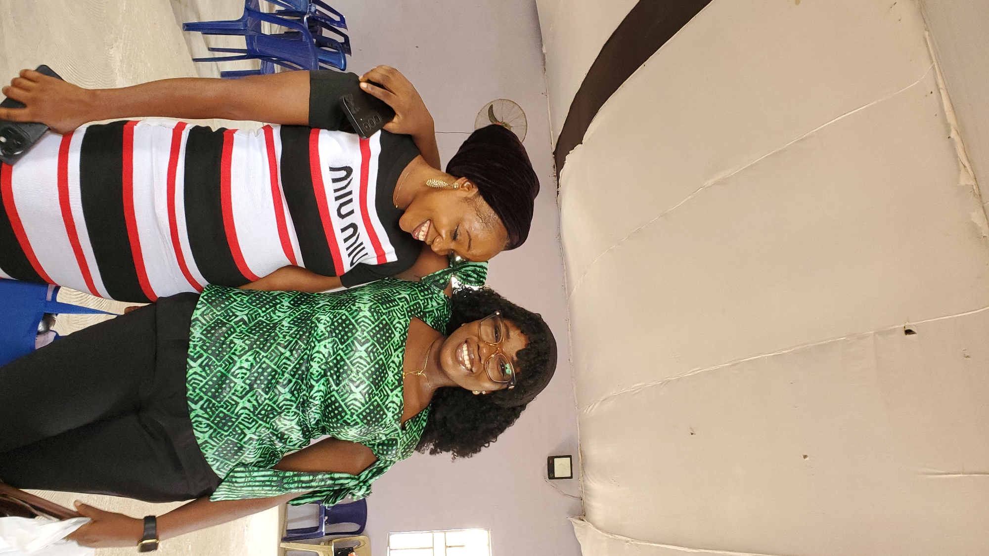The image depicts two women standing indoors, possibly in a community hall or event space in Jalingo, Nigeria. The woman on the left wears a black headwrap, gold earrings, and a striped dress with black, white, and red horizontal bands. She is holding a smartphone in her left hand and has her right arm around the shoulder of the other woman. The woman on the right has curly dark hair, wears glasses, and a green patterned top with tie details on the sleeves. She is also wearing black pants and a watch on her left wrist. Both women are smiling and appear to be posing for a photo. In the background, blue plastic chairs are visible, suggesting a gathering or event. The ceiling is a suspended white fabric with visible seams and some damage. A wall fan is mounted on the back wall, and a light fixture is also present.