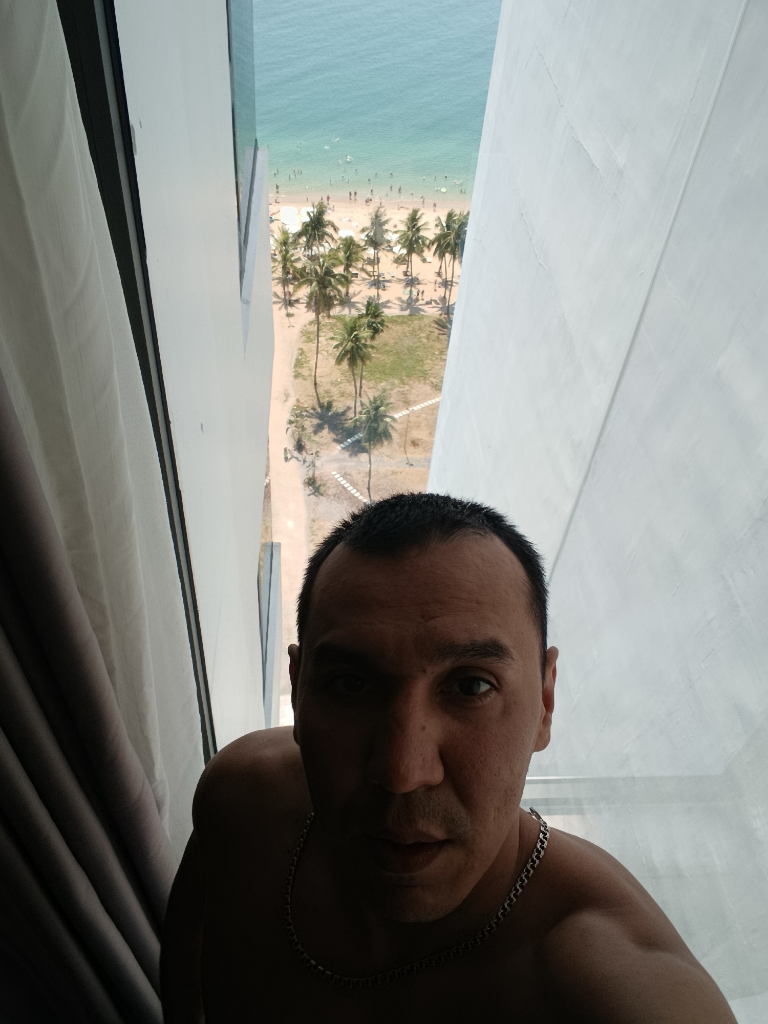 A medium-closeup shot from an elevated perspective showing a partially obscured man, likely in a hotel room, looking into the camera. Behind the man is a narrow view of a beach with palm trees, people, and the ocean, located in Nha Trang Ward, Vietnam. The man is wearing a silver chain around his neck and appears to be shirtless. The framing suggests he is standing next to a window or balcony. The lighting is uneven, with the man partially in shadow.