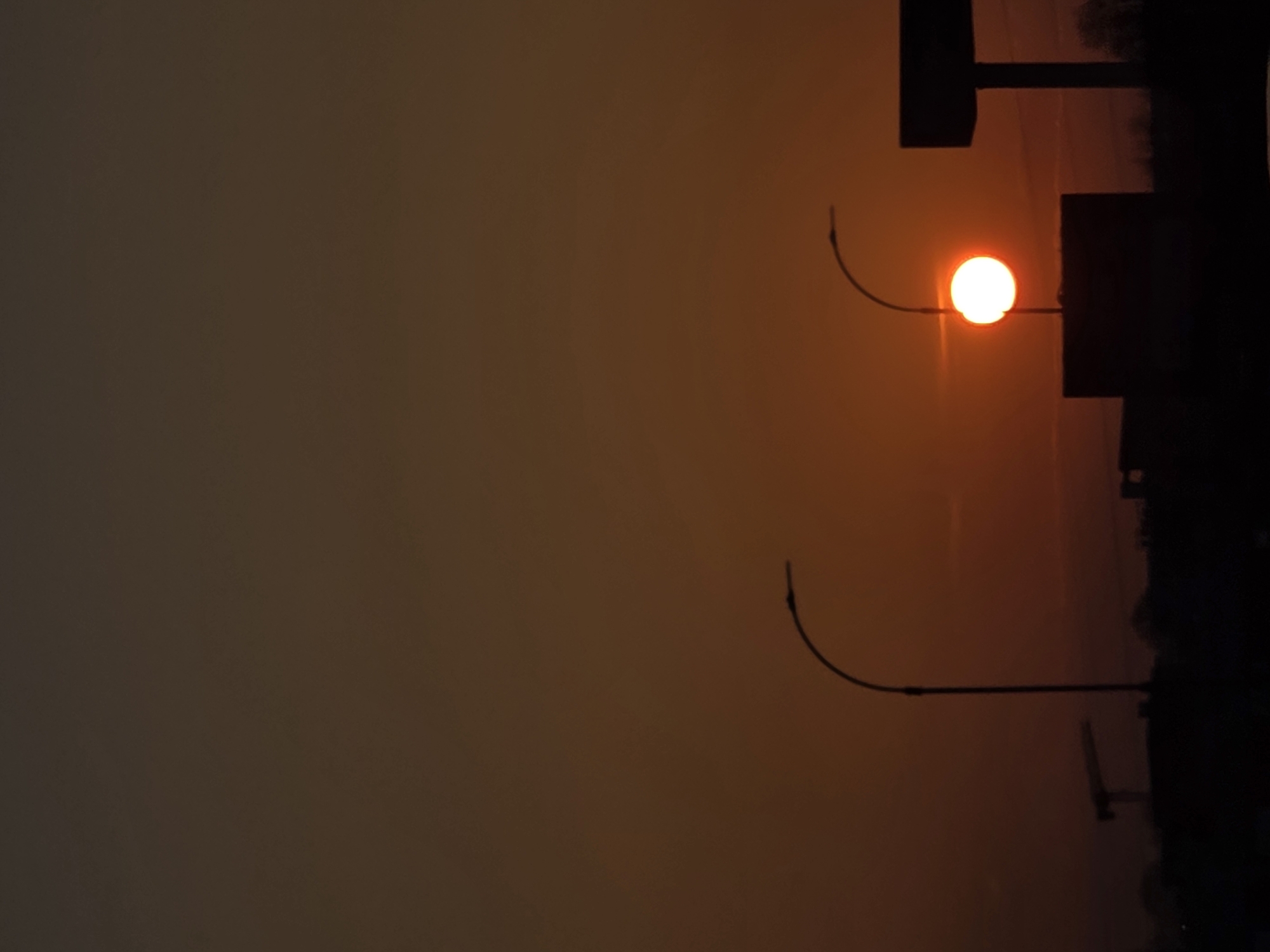 The image depicts an urban or industrial periphery scene in Varna, Bulgaria, at either sunrise or sunset. The sky presents a gradient of warm hues, transitioning from deep orange-red at the horizon line to a darker brown-orange overhead. A prominent, bright white-yellow sun disc is positioned slightly right of the frame's center, partially obscured by the upper edge of a dark, rectangular building silhouetted against the sky. Two streetlights with tall, curved poles are visible in silhouette: one directly to the left of the central building, and another smaller one further left. On the far left horizon, the dark outline of a construction crane is discernible. To the right of the central building, a large, T-shaped structure, consistent with a billboard or sign support, rises prominently in silhouette. Minimal sparse vegetation outlines are faintly visible on the far right edge of the horizon. No individuals or dynamic actions are present within the frame.