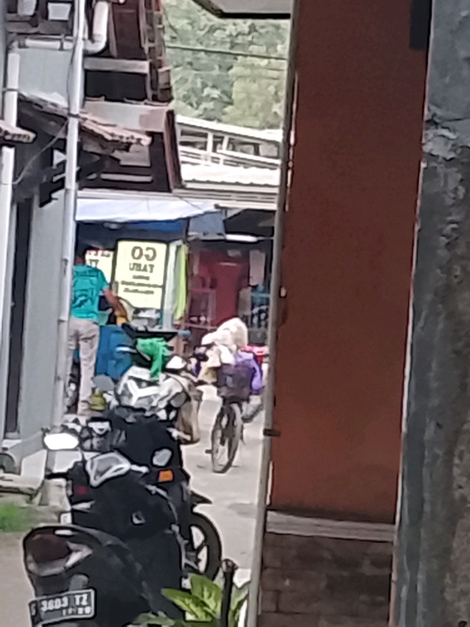 In Ujungrusi, Indonesia, a man in a turquoise t-shirt and light-colored pants is standing in front of a storefront. He is holding a bag or sack and appears to be interacting with items inside the store, which has a yellow sign with black text that reads "CO UBAT." Behind him, a bicycle with a basket full of items, including what appears to be white cloth, is parked. In the foreground, a black motorcycle with a visible license plate that reads "S 3603 TZ" is parked. The environment suggests a street scene with buildings and vegetation visible in the background.