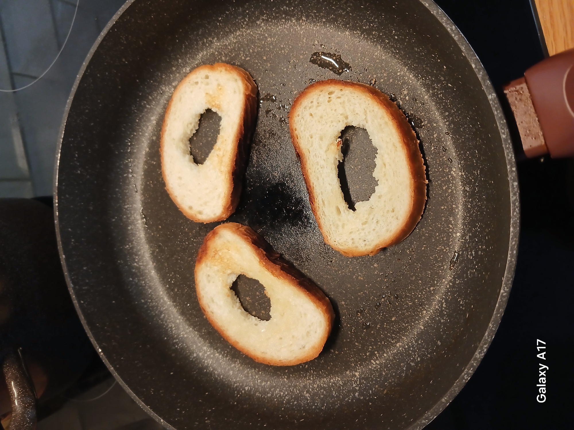 A top-down, close-up shot depicts three pieces of bread, each with a circular hole in the center, frying in a black non-stick pan with a brown handle. The bread is lightly toasted with a golden-brown crust. A small amount of oil is visible in the pan. The pan rests on a surface, possibly a stove, with a partial view of what appears to be a cooktop in the upper left. The bottom right of the image includes the text "Galaxy A17". There is no indication of the city of Yekaterinburg, Russia, or any other identifying information about the location.