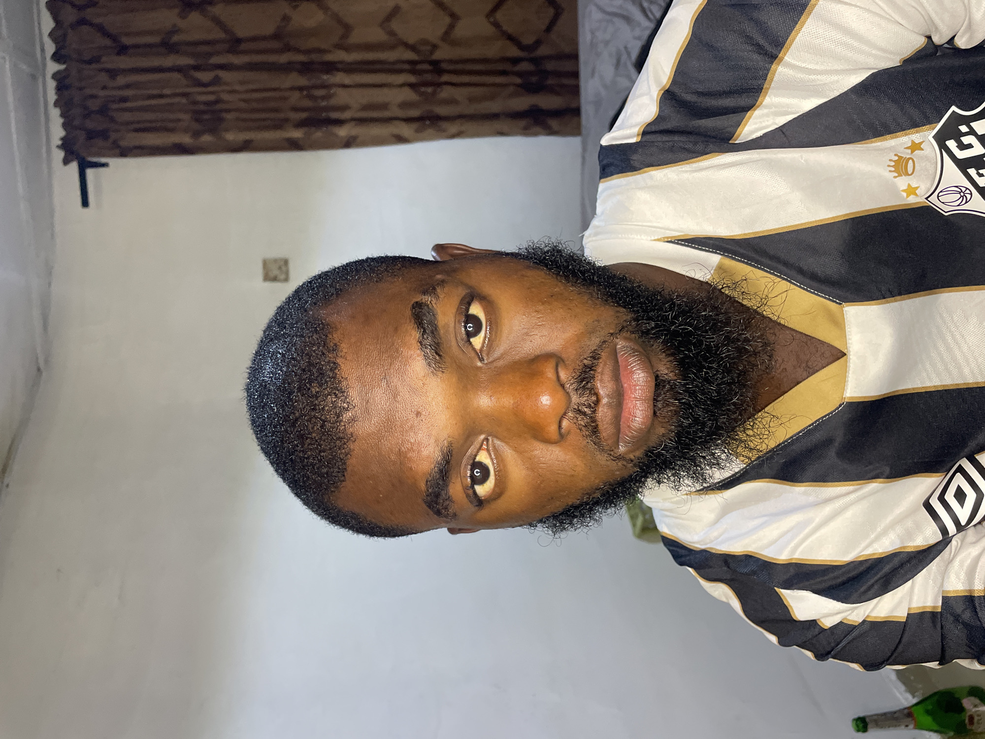 The image shows a close-up portrait of a Black male from the chest up, looking directly at the camera. He has dark, short hair and a full beard and mustache. He is wearing a black and white striped jersey with gold trim. The jersey has a crest on the left side that appears to be a sports team logo, featuring crowns, stars, and a basketball icon. The background is blurred, showing a plain wall with a brown curtain on the right side and a small square decoration on the wall. A bottle with a green label is partially visible on the left side of the frame. The lighting appears to be artificial, coming from the front and slightly from the left, creating some shadows on his face. The overall scene suggests an indoor setting. There is no information in the image to indicate the location is the city of Jos, Nigeria.