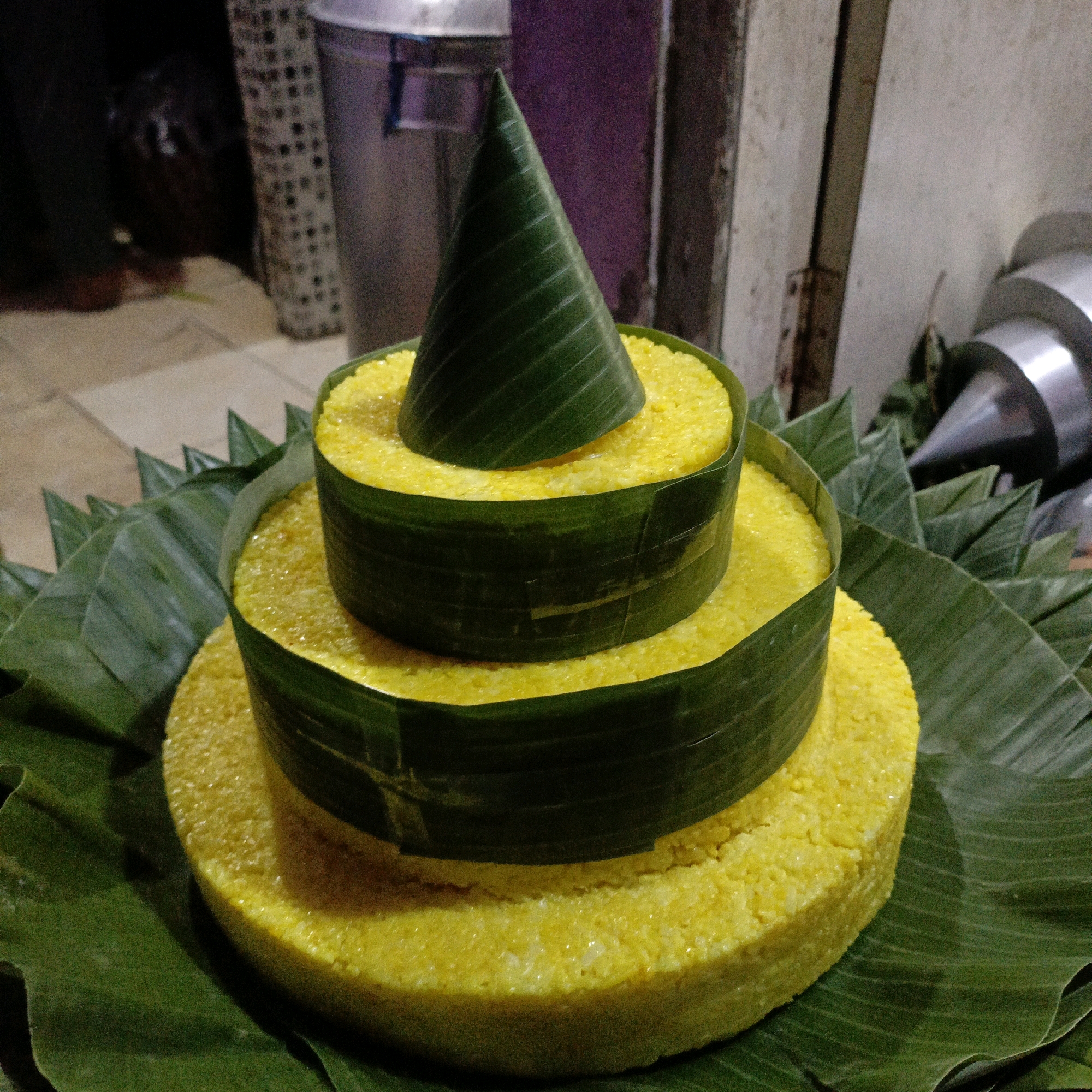 In Salatiga, Indonesia, a tiered, yellow rice cake, likely a traditional delicacy, is presented on a bed of large, green banana leaves. The cake is arranged in three cylindrical layers, each encircled by a band of banana leaf. The top layer is crowned with a cone-shaped structure also made from a banana leaf. The background is dimly lit and shows hints of urban elements, including a large, metallic trash can to the left and what appears to be a metal funnel or spout to the right. The overall setting suggests an outdoor market or street stall environment typical of Salatiga.