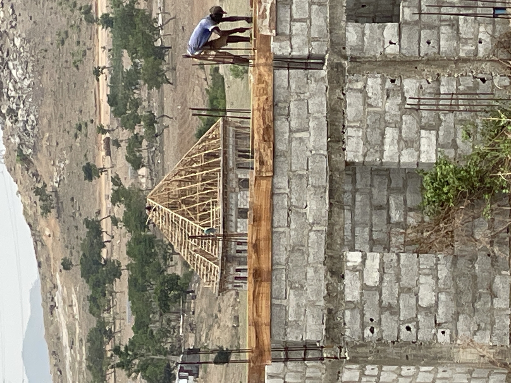 A medium-shot captures an ongoing construction site in Angwan Sayawa, Nigeria, displaying the partial construction of a building with a gray block wall structure. A worker sits atop a makeshift platform, positioned against the wall. To the left, the wooden framework of a roof structure is visible. The background reveals an arid landscape with a sparsely vegetated hillside.