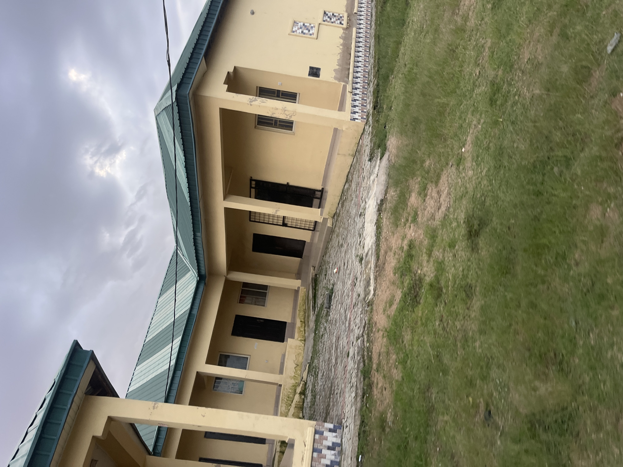 The image depicts a single-story building with a green corrugated metal roof and light yellow walls, located in Jos, Nigeria. The building features a covered walkway supported by pillars, with several doorways and windows visible. The ground in front of the building is paved with interlocking bricks, and an area of green grass is present to the left and foreground. The sky is overcast with dramatic clouds. The building's exterior shows some signs of wear and tear, with peeling paint and some discoloration. Decorative tiles are visible along the lower portion of the building. There are no people or animals visible in the image. The scene appears to be captured during daylight, under what seems to be an approaching storm given the dark clouds.