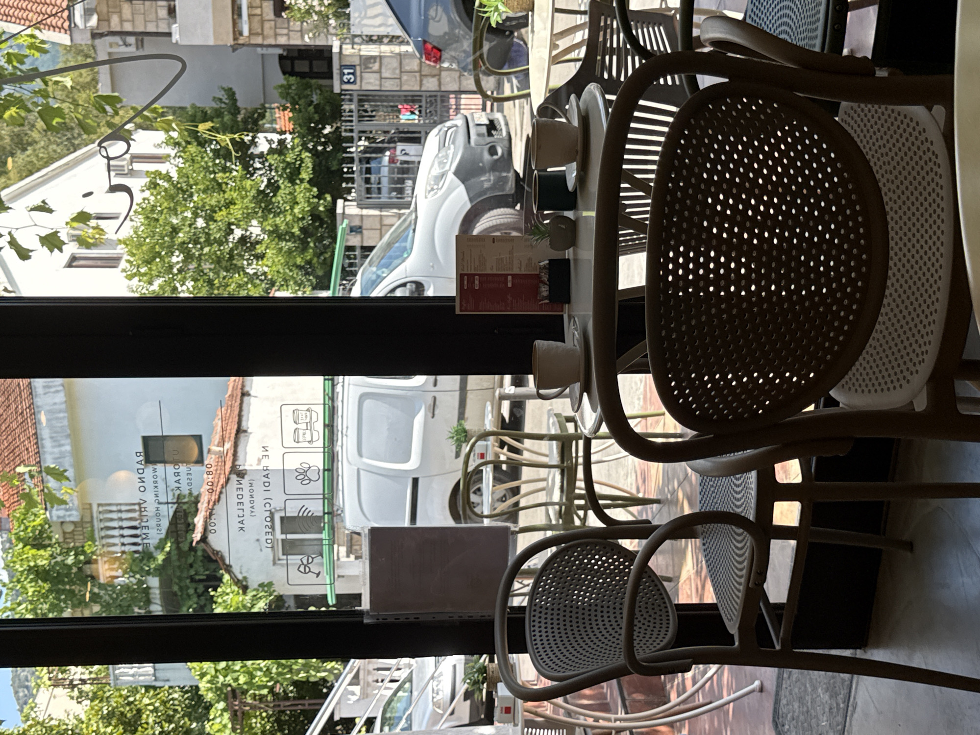 The image shows the outdoor seating area of a cafe in Budva, Montenegro, as viewed through a glass window. In the foreground, several chairs and small tables are visible. Two of the chairs are modern, made of a woven material with a metal frame. There are two coffee cups and saucers on one of the tables, along with a small potted plant and a sugar dispenser.
Through the glass, a street scene is visible. A white van is parked on the street, and a grey car is parked further down. Buildings with red-tiled roofs and white facades line the street, with lush green trees and foliage providing a natural backdrop. A sign on the window displays operating hours in a mirrored format, indicating "OPEN 08:00 TUESDAY - SUNDAY" and "CLOSED MONDAY." The overall impression is a bright, sunny day in a European coastal town.