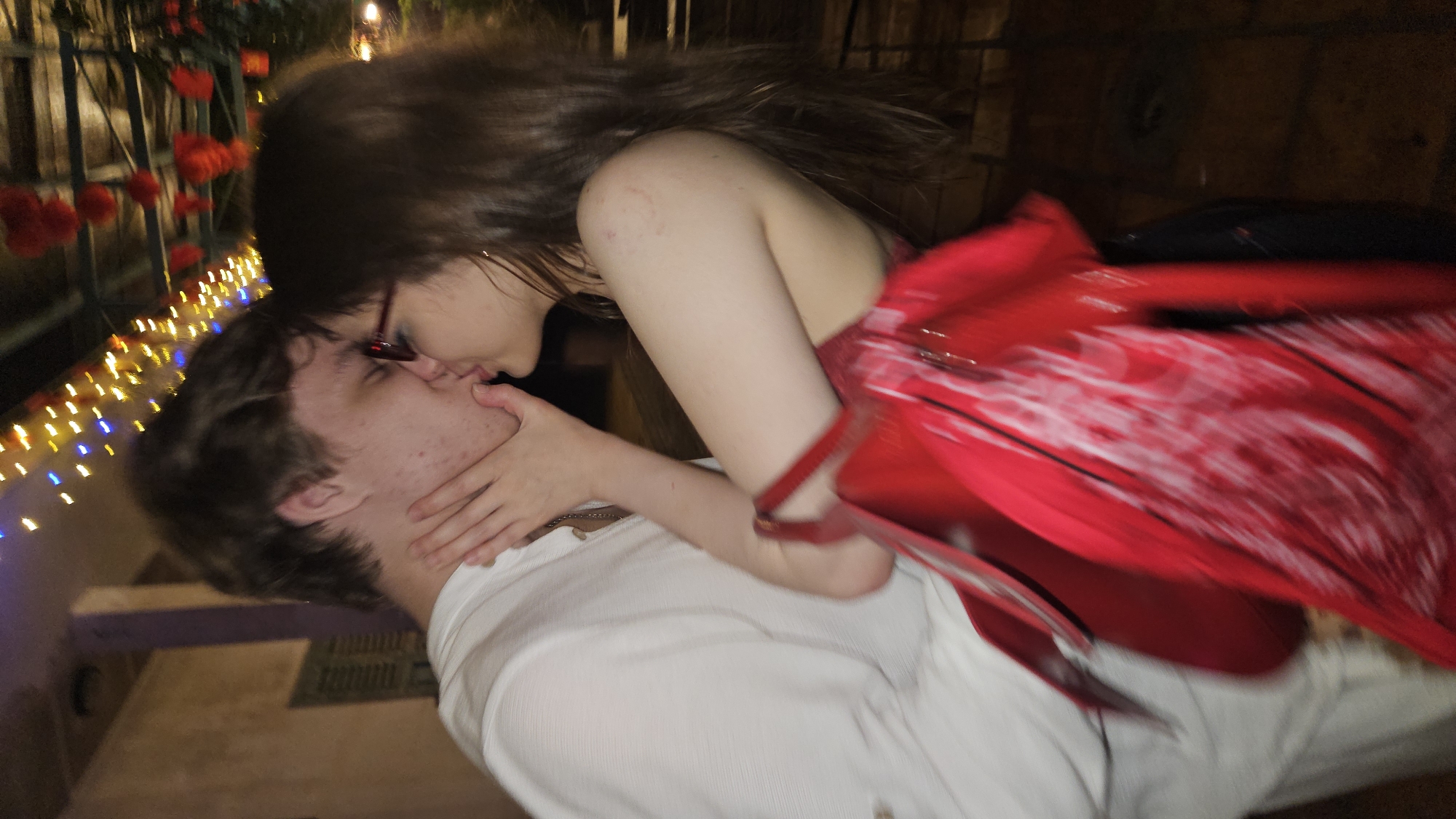 A couple is kissing. The man is wearing a white shirt and is lying down. The woman is wearing a red dress and has dark hair. She is wearing glasses. The background includes string lights and what appears to be a building facade. The location cannot be identified as Nha Trang, Vietnam from the image alone.
