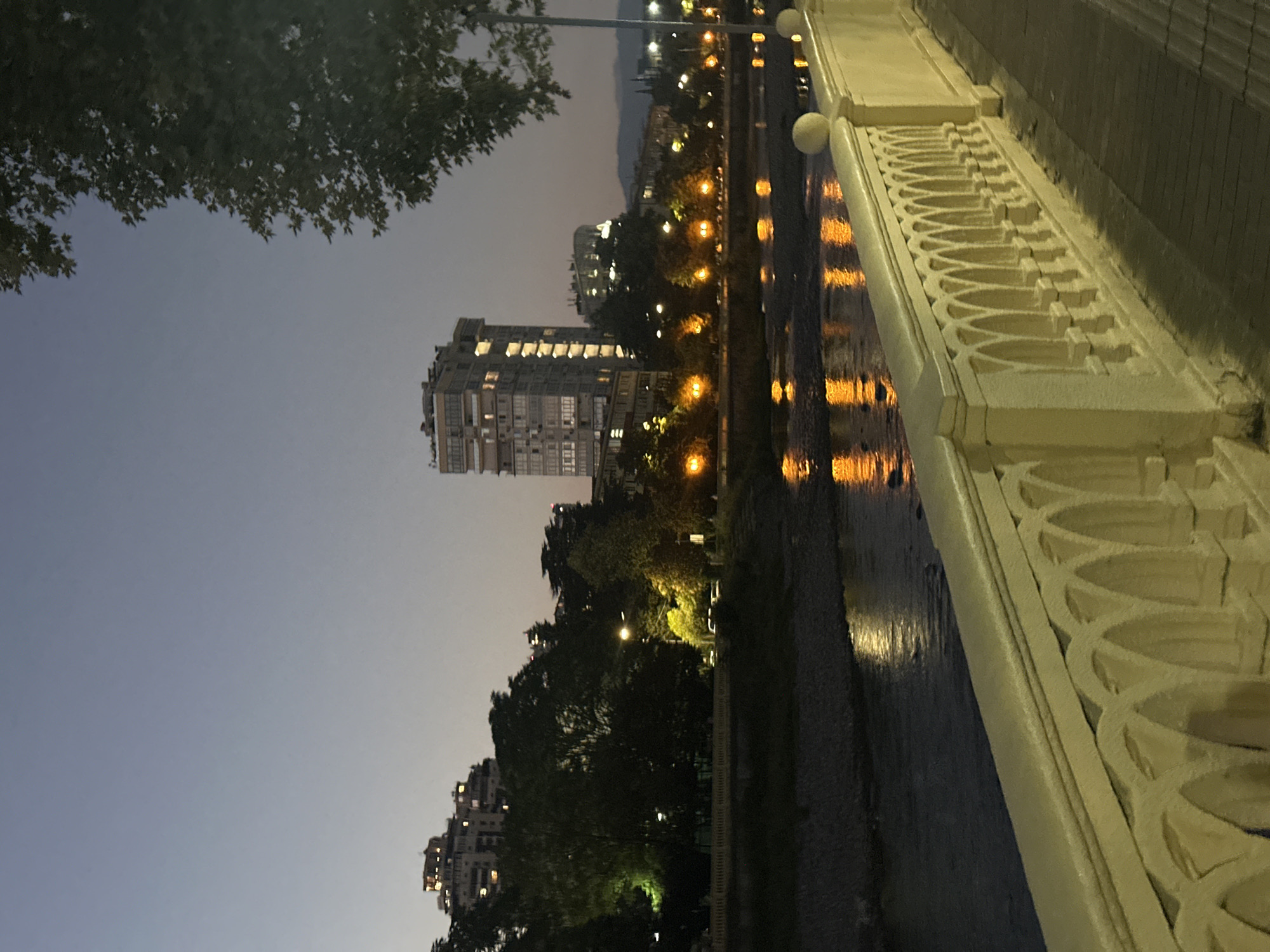 The image depicts a nighttime scene along a riverfront promenade in Sochi, Russia. A cream-colored ornate balustrade runs along the right foreground, with arched detailing. Beyond the balustrade, a paved walkway is visible. The river flows to the left of the walkway, reflecting the warm glow of streetlights and building lights.
Across the river, a tree-lined embankment is visible, dotted with illuminated street lamps casting golden pools of light. In the background, several buildings are illuminated, including a tall, modern high-rise with many visible windows and balconies. Further in the distance, more buildings and the silhouette of hills can be seen against the twilight sky. A large tree with dense foliage partially obscures the upper right portion of the image. The overall atmosphere is calm and serene, characteristic of a city at dusk or early evening.