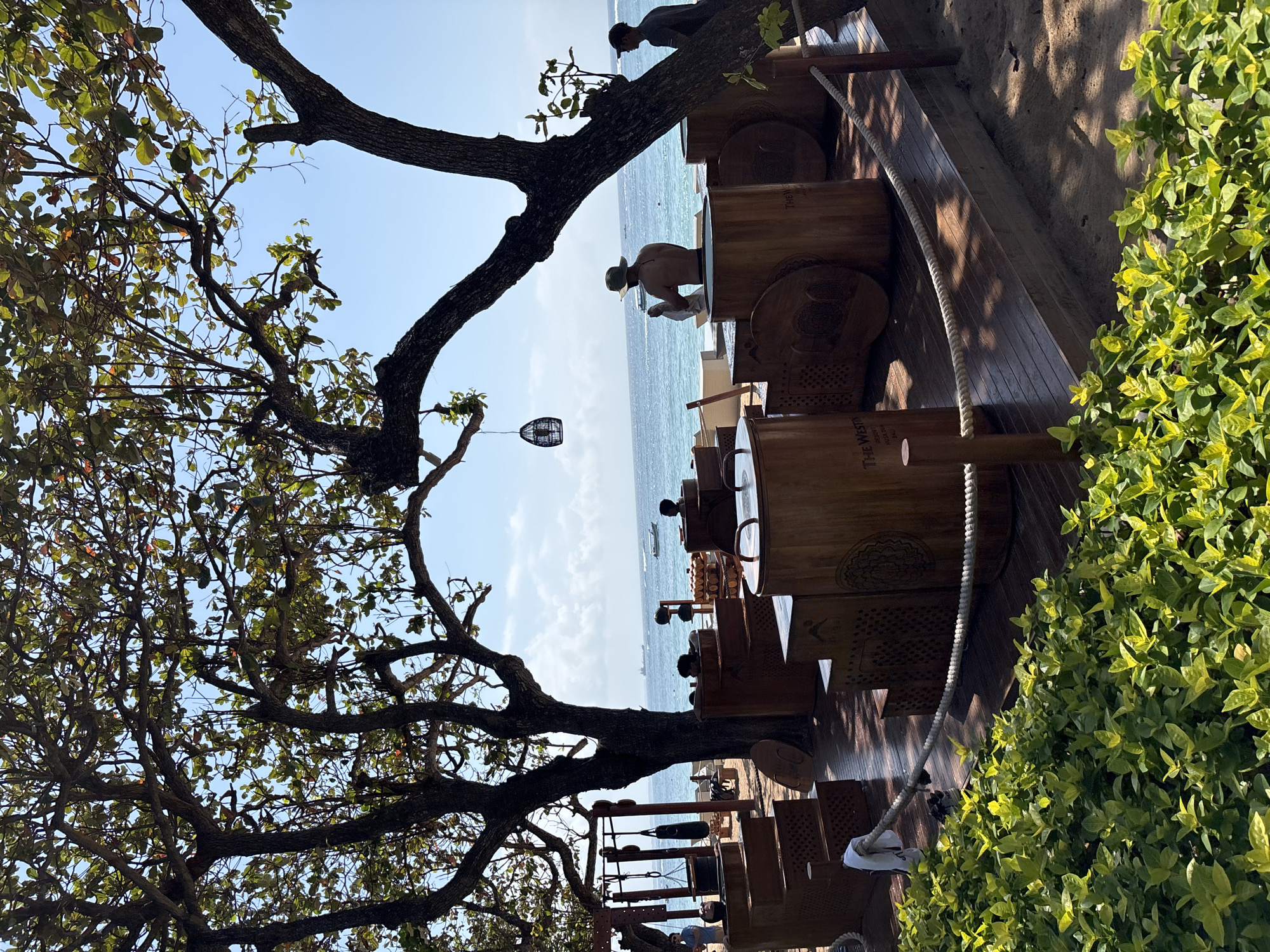 The image shows an outdoor seating area by the sea in Kuta Selatan, Indonesia. In the foreground, a lush green hedge lines the bottom of the frame. Behind the hedge is a wooden deck with several large, cylindrical wooden structures, some with seating areas. A thick rope barrier runs along the edge of the deck.
In the middle ground, under the shade of large trees, a man wearing a hat is sitting in one of the wooden structures, holding a white towel. Another man is standing to his right. In the distance, the turquoise ocean stretches to the horizon, with a few boats and ships visible. The sky is a clear blue with scattered white clouds.
To the left of the wooden structures, there is workout equipment, including punching bags hanging from a frame. The overall scene suggests a resort or spa area, likely on a beach.