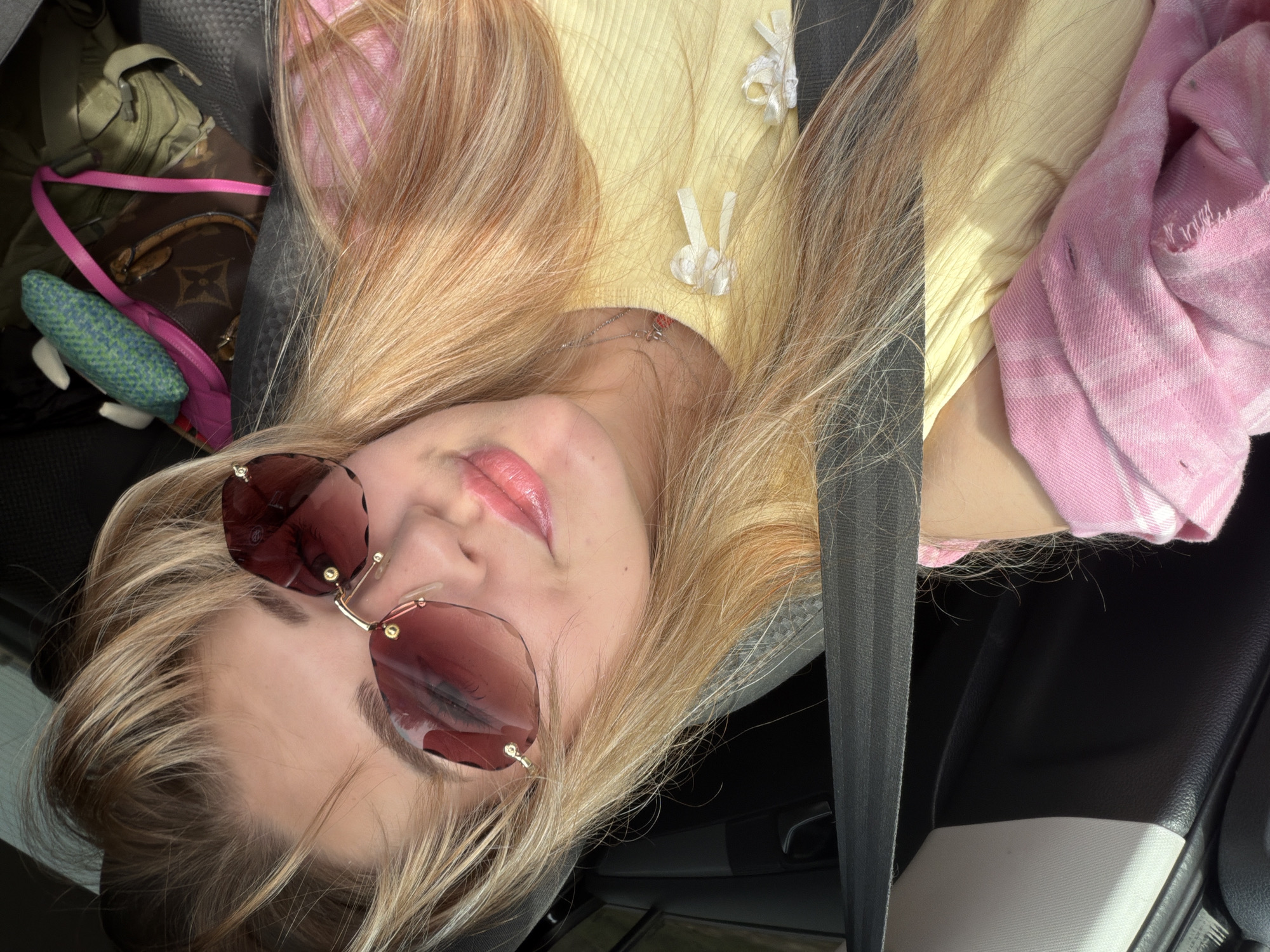 A young woman is reclining in what appears to be a car. She is wearing sunglasses and a yellow top with decorative ties. A pink and white patterned garment is visible to her right. A black seatbelt crosses diagonally in front of her. There is a bag with a brown and tan pattern, a pink strap, and a green and blue stuffed object in the background. The interior of the car is visible, with black and gray surfaces. I can't find any relevant context about a city called Псеево.