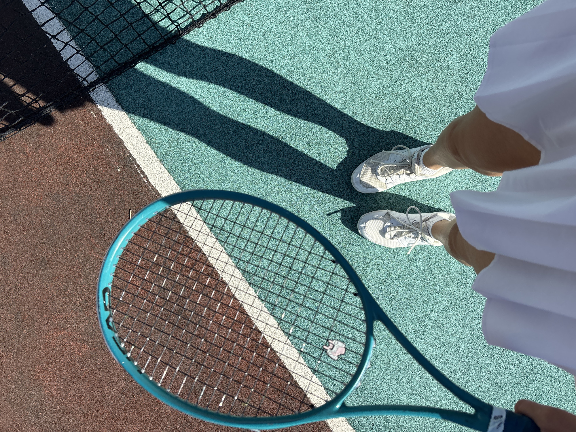 The image shows a first-person perspective of a person on a tennis court. The court has two distinct colored surfaces: a reddish-brown area and a teal-colored area, separated by a white line. A portion of a tennis net is visible in the upper right corner. The person is holding a teal-colored tennis racket in their left hand and is wearing white tennis shoes and a white skirt. The person's legs and feet are visible. The scene is outdoors, as evidenced by the shadows cast on the court, indicating a sunny day. The image was taken in the city of Chaikovsky, Russia, specifically at a tennis court located at ulitsa Vinnitskaya, 38.