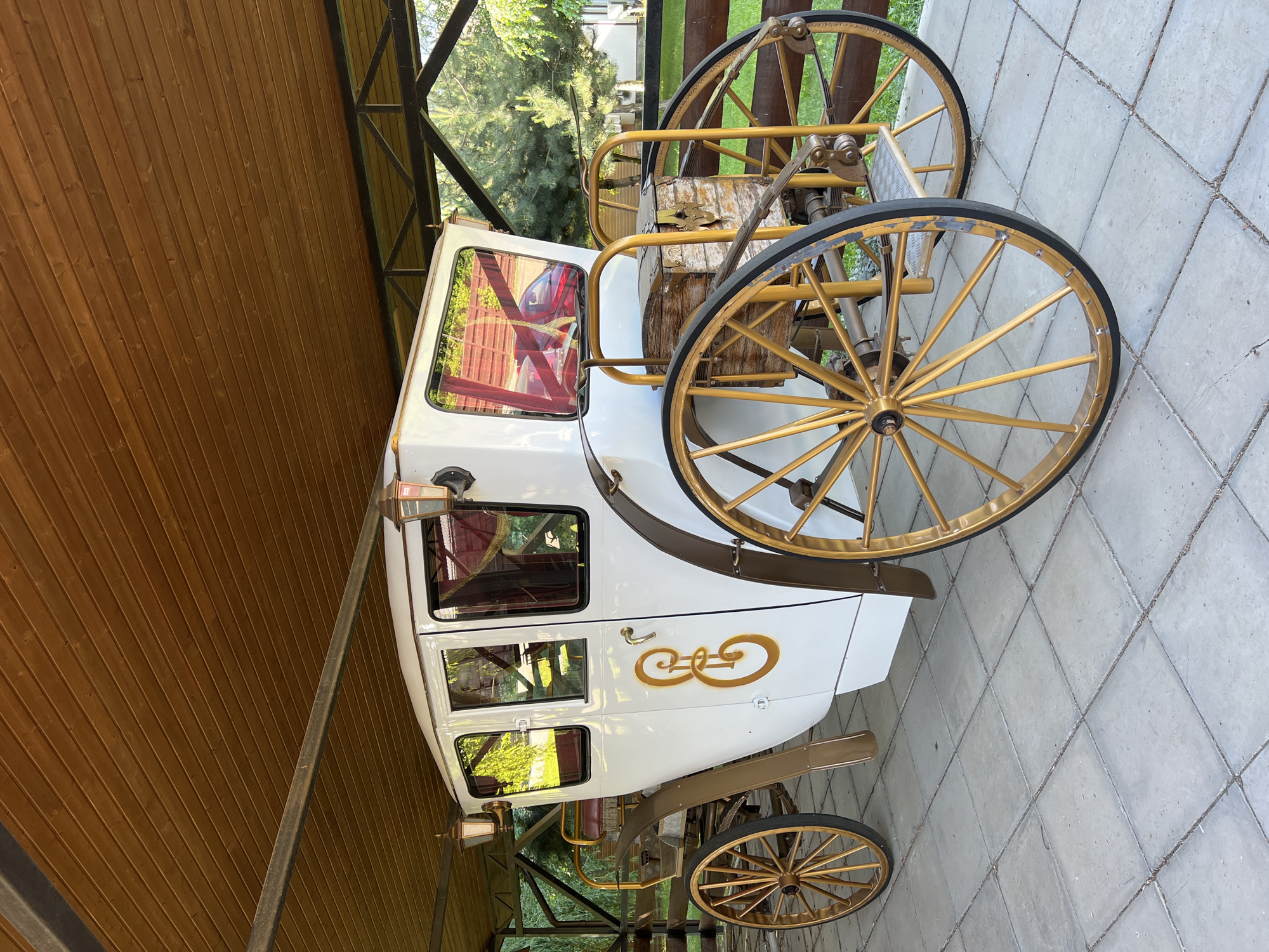 A white carriage with golden trim and wheels is parked under a wooden canopy. The carriage has large windows and intricate golden lettering on its side. The wheels are large and spoked, with black tires. Behind the carriage, a wooden fence and trees are visible. The ground is paved with square tiles. There are no people visible in the image.