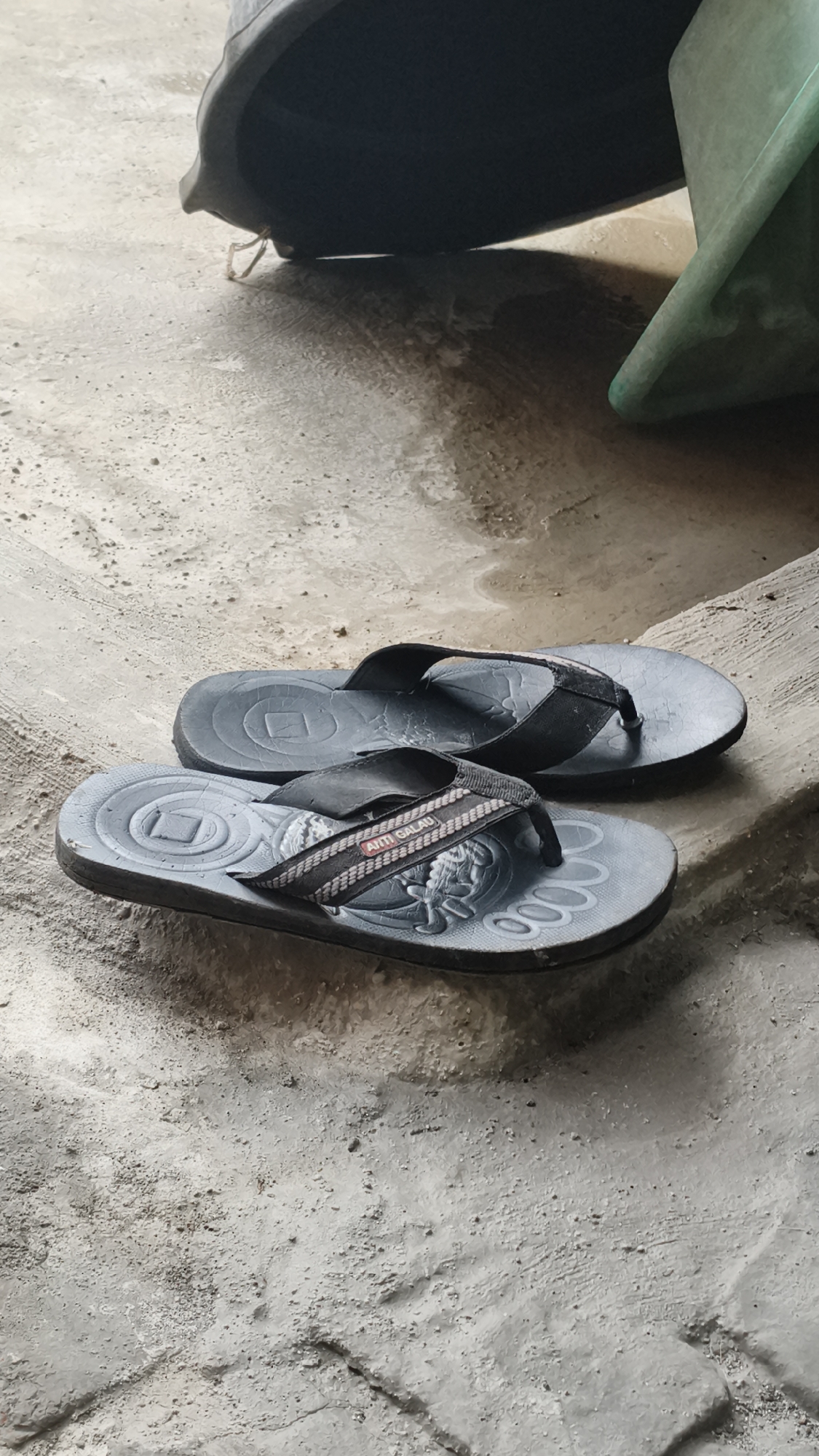 A pair of black flip-flops rests on a concrete surface. The flip-flops are oriented horizontally, with the right flip-flop slightly in front of and to the right of the left flip-flop. Both flip-flops appear to be worn, with some scuff marks and slight fading. The straps are black with a textured pattern, and one strap has a label that reads "ANTI GALAU." The concrete surface shows signs of wear, with some cracks and unevenness. In the background, out of focus, are parts of dark-colored and green objects, possibly buckets or containers. The scene is outdoors, as indicated by the concrete and the lighting. This image was taken on a street in Kerep Kidul, Indonesia.