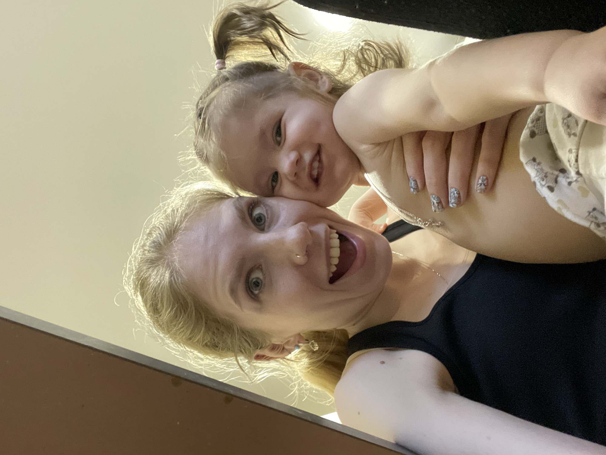 The image shows a selfie of a light-skinned woman with blonde hair, wearing a black tank top. She is holding a young child with light skin and blonde hair pulled back in a ponytail with a pink hair tie. Both are smiling at the camera. The woman has a nose ring and is wearing a silver necklace. The child is wearing a light-colored garment. A partially visible brown object is present in the lower left corner of the frame and a dark object is present in the upper right corner. The background appears to be a light tan wall.
It is not possible to identify the location as Izhevsk, Russia or any other specific location.