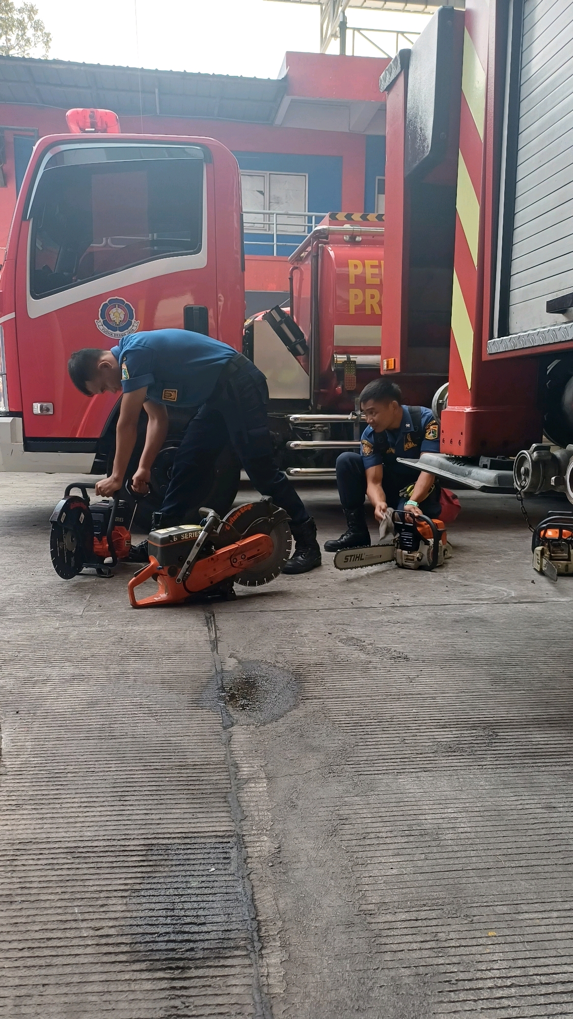 Two firefighters in blue uniforms are working on the ground in front of a red fire truck. The firefighter on the left is bending over two large orange cutting tools, possibly concrete saws, with one hand on the handle of a saw. The firefighter on the right is kneeling and holding a smaller chainsaw, with a cleaning cloth in his other hand. Both individuals are wearing black boots. The ground is a gray concrete surface with distinct parallel lines and some wet patches. In the background, part of the red fire truck is visible, along with a building with blue walls and white-framed windows. The scene appears to be within a fire station, likely in South Jakarta, Indonesia, based on the context of the fire truck and uniforms.