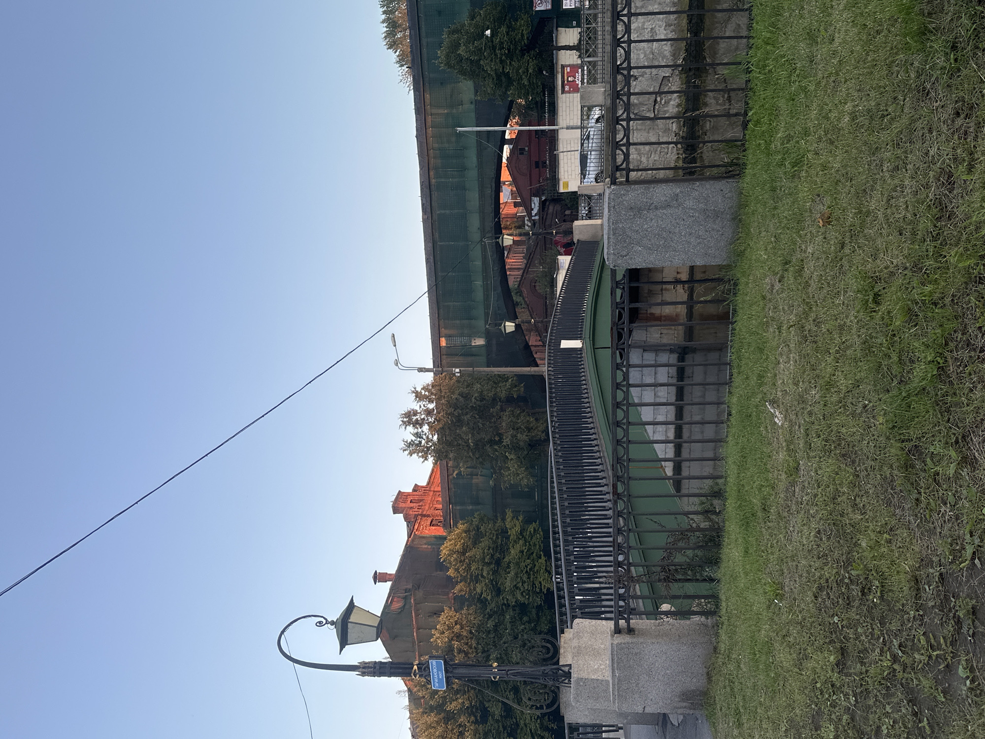 A view of the Tarakanovsky Bridge in Saint Petersburg, Russia. The bridge is a pedestrian bridge, painted green, with black iron railings. It spans a canal. In the background, a larger, green-painted railway bridge crosses over the canal. Red brick buildings with ornate architecture are visible under the railway bridge, some appearing to be under renovation with scaffolding and green netting. A vintage street lamp with a decorative post stands on the left side of the image. A blue street sign with white Cyrillic text is attached to the lamp post. The scene is bathed in the warm light of either sunrise or sunset, casting orange hues on some of the buildings. The foreground consists of a grassy area with sparse vegetation. The sky is clear and blue.
