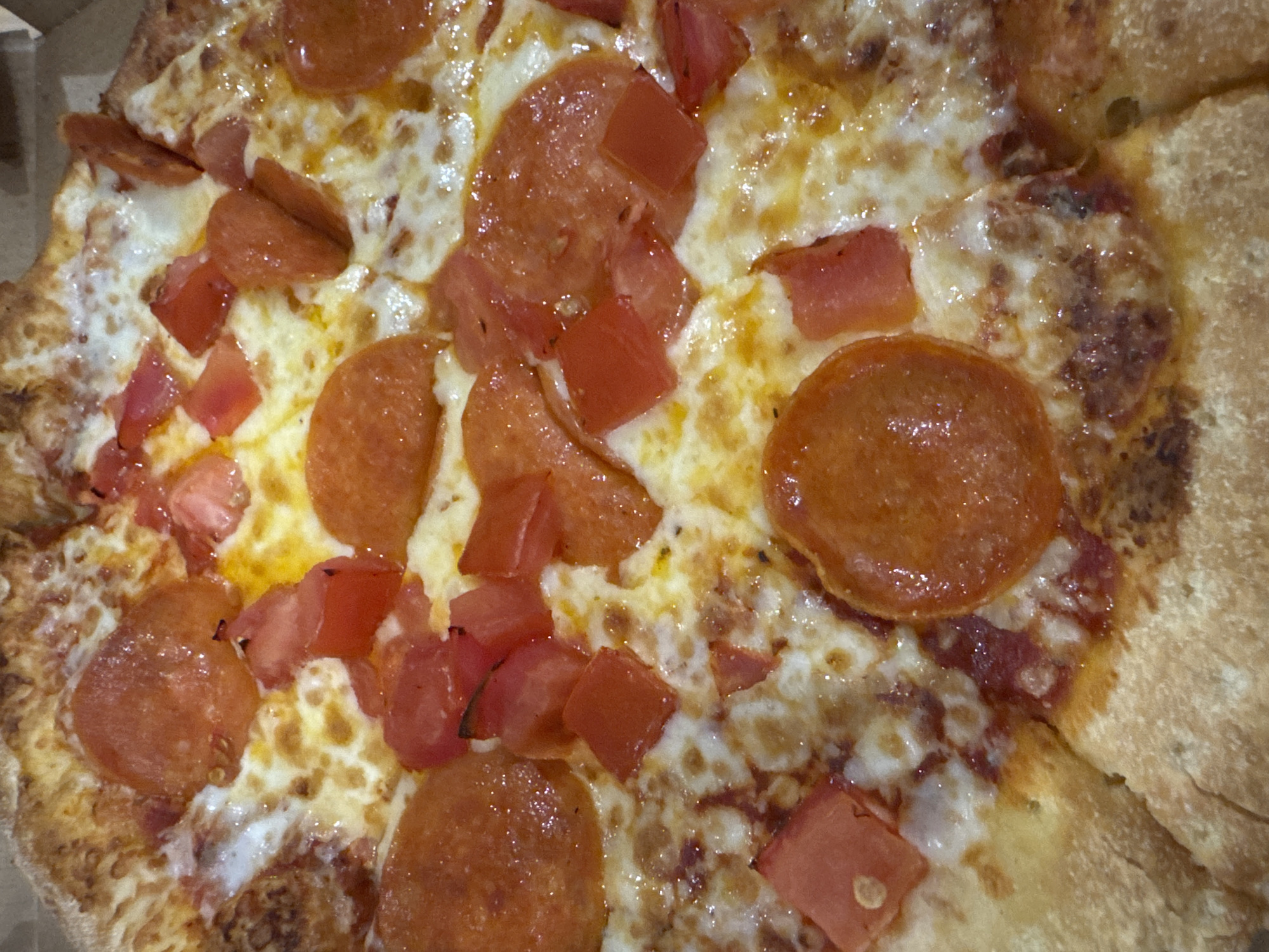 A close-up, overhead view depicts a pizza resting within a light brown cardboard box, visible along the top right edge. The pizza is generously covered with melted, bubbly white and yellow cheese, which displays an oily sheen. Numerous circular slices of cooked pepperoni, orange-red in color, are interspersed across the cheese. Alongside the pepperoni are irregularly diced pieces of bright red tomatoes. The pizza crust, visible at the bottom and along the periphery, is golden brown and appears slightly textured. No individuals, specific actions, or further environmental details are present. The scene is located in Votkinsk, Russia.