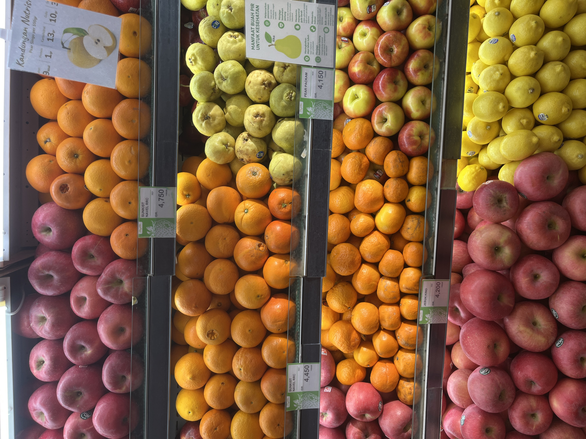 The image shows produce for sale in a North Kuta, Indonesia market. The produce includes apples, oranges, pears, and lemons, separated in sections. Nutritional information is displayed on a sign for the pears. Prices are also posted.