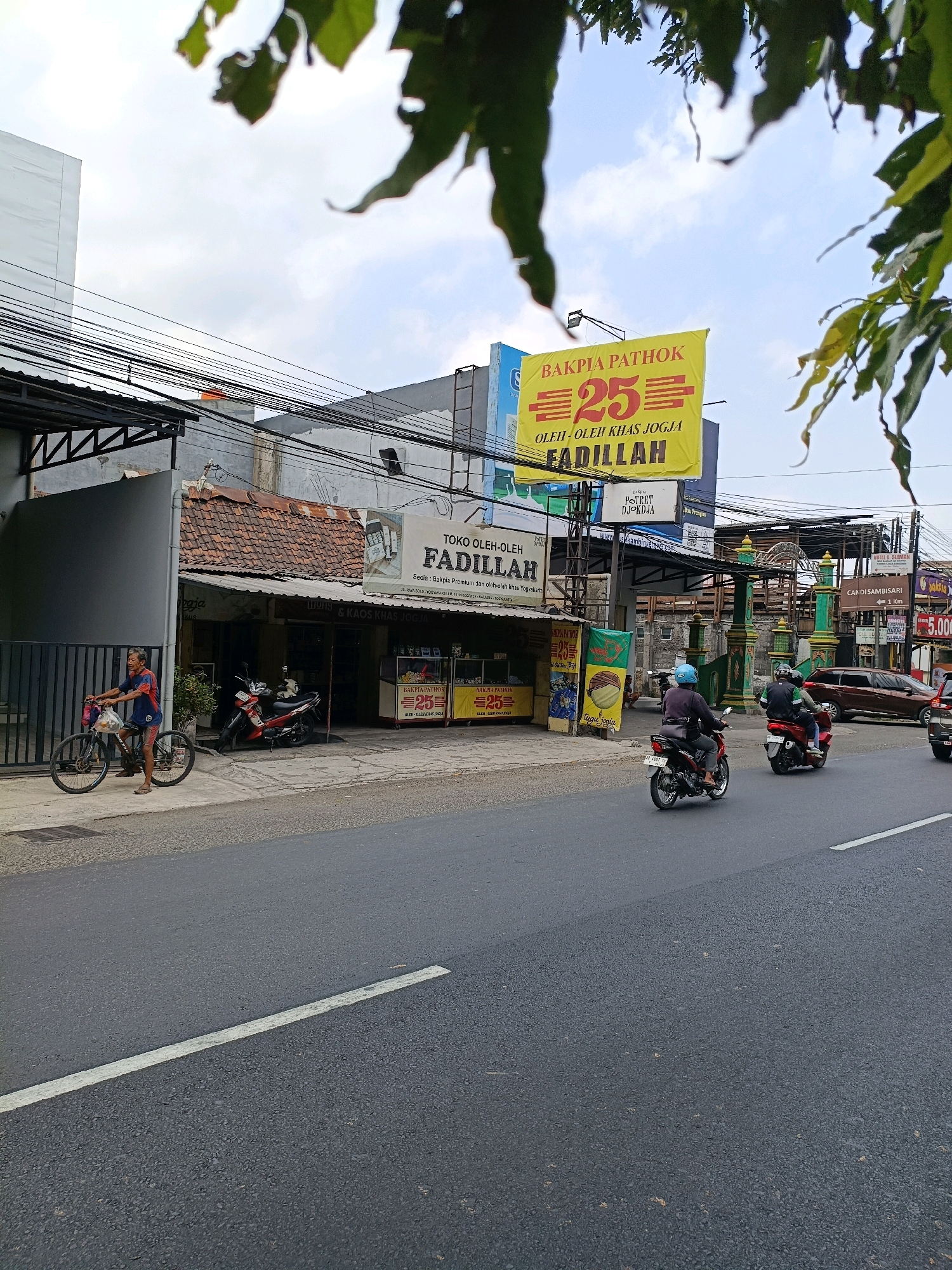 In Purwomartani, Indonesia, on a cloudy day, a man rides a bicycle on the sidewalk of a street. He is wearing a blue shirt and red shorts. In the background, there is a building with a tiled roof and a storefront with a sign that reads "TOKO OLEH-OLEH FADILLAH". Another sign above the storefront advertises "BAKPIA PATHOK 25". Several motorcycles are parked in front of the storefront. Further down the street, two people are riding a red motorcycle, and a brown SUV is driving on the road. The street is paved, and there are power lines overhead. The scene depicts a typical street view in a bustling commercial area, with signs for local businesses and traffic on the road.