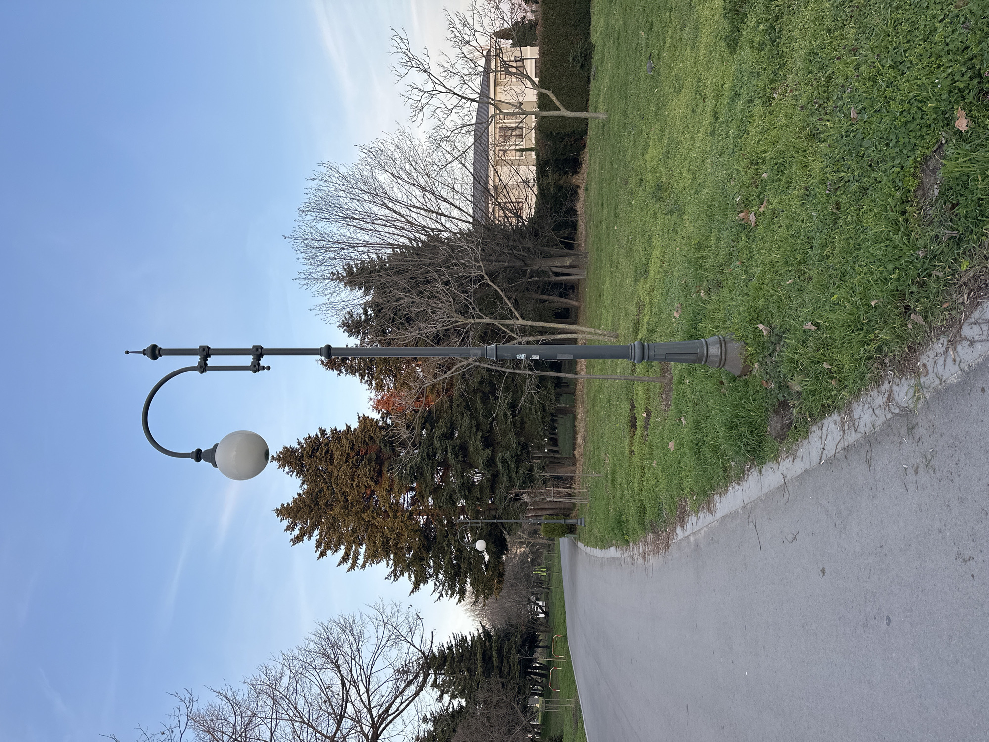 The image depicts a park setting, likely in Varna, Bulgaria, although specific landmarks are not visible to confirm this location. A paved pathway cuts through the scene from the foreground towards the background. To the right of the path is a grassy area with a mixture of grass and other ground cover. On the left side of the path, a row of trees lines the edge. A prominent lamp post with a spherical globe is positioned close to the left edge of the path. In the background, a building is partially visible behind the trees. The sky is blue with wispy clouds. There is no indication of any people or activity in this image.