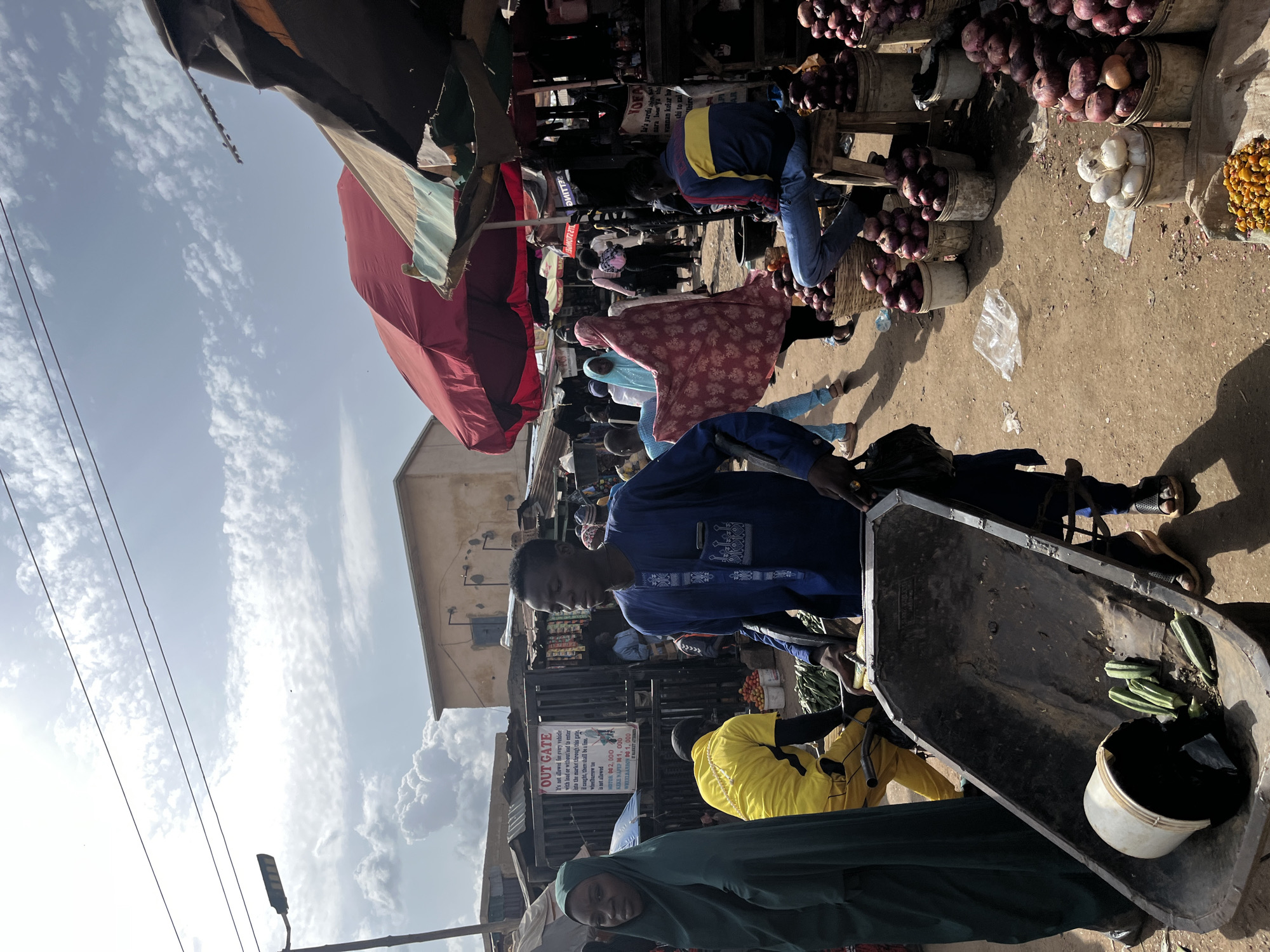 The image depicts a busy outdoor market scene in Rafin Par, Nigeria. In the foreground, a man in a blue traditional outfit pushes a large, weathered wheelbarrow containing a white bucket, some black plastic bags, and several green vegetables, possibly okra. To his left, a woman in a dark green hijab is partially visible. Behind them, a man in a bright yellow shirt is seen on a bicycle.
The market is lined with stalls covered by awnings and red umbrellas, displaying various goods. Piles of red onions are prominently displayed in baskets and buckets on the right side of the image. In the background, other people are seen moving through the market, some dressed in colorful clothing. A sign above a stall reads "OUT GATE" with additional text in what appears to be Hausa, indicating market regulations and possibly prices. The ground is dusty, and the sky above is partly cloudy with bright sunlight. The overall atmosphere suggests a bustling commercial hub.