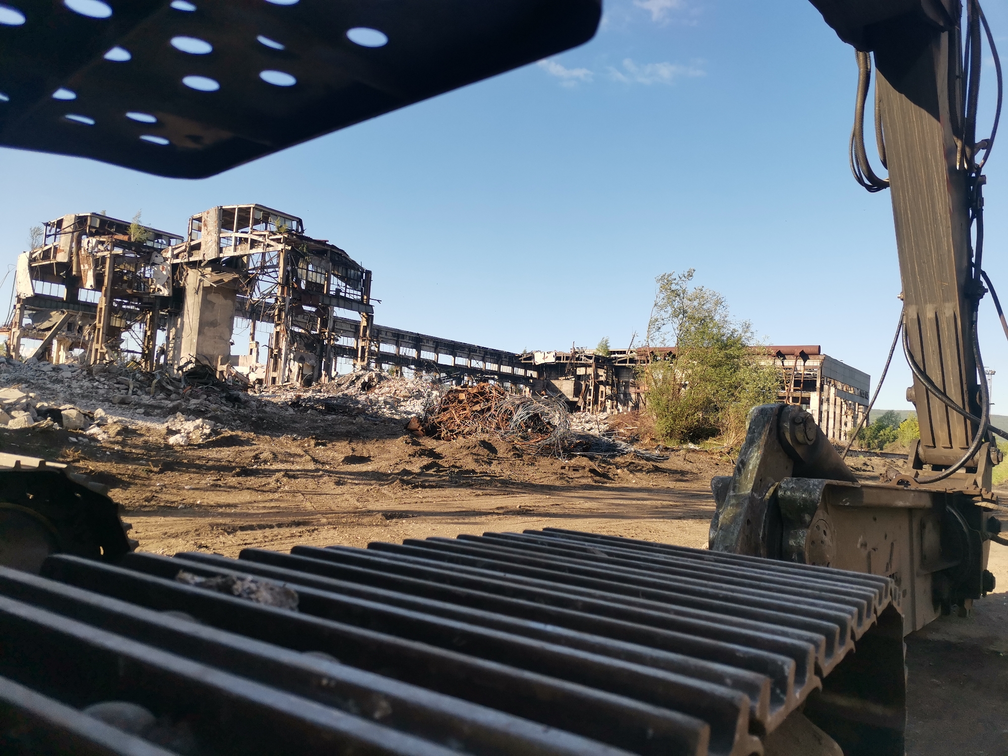The image shows the skeletal remains of a large industrial building in Zhivkovo, Bulgaria. Debris and piles of scrap metal litter the ground. A piece of heavy machinery, possibly an excavator or crane, is prominently positioned in the foreground, with parts of its tracks and arm visible. The machine is facing the building, suggesting it may be involved in demolition or cleanup. The sky is clear and blue.