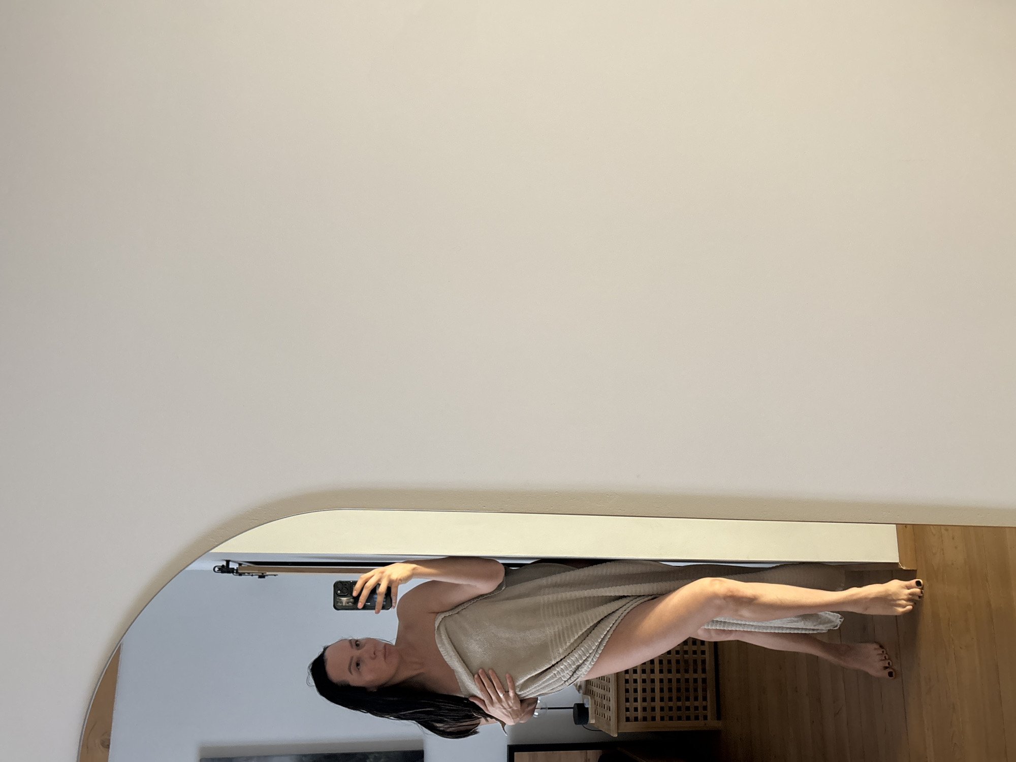 A woman, draped in a beige towel, is taking a selfie in front of a round mirror. She holds her phone in her left hand, capturing her reflection. Behind her, the room includes a white wall, light-colored wood floor, and glimpses of furniture, including a cabinet. The location of the image is not explicitly stated.