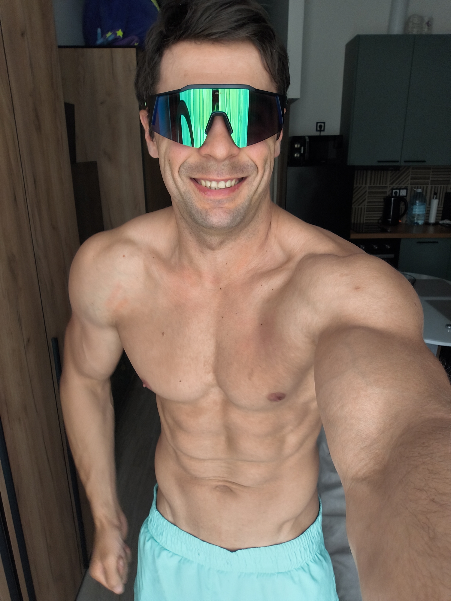 A muscular man with short dark hair and wearing reflective green sunglasses is seen from the chest up, smiling and facing the camera. He is shirtless and wearing light blue shorts. The background appears to be a residential interior, with a wooden wardrobe to the left and kitchen cabinets and appliances visible behind him. There is no information in the image to indicate that this photo was taken in Moscow, Russia.