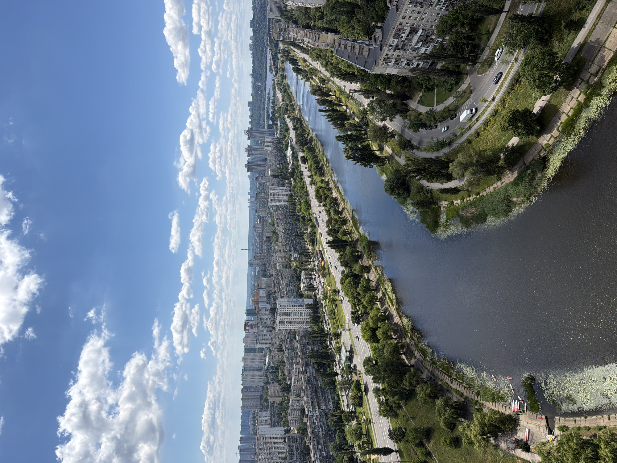 This is an aerial, vertically oriented photograph of a city skyline on a clear, sunny day. The scene depicts a wide, dark river running through a densely populated urban area characterized by numerous apartment buildings and trees. A road with visible traffic, including cars and a truck, runs alongside the river, lined with more trees and pedestrian walkways. In the foreground of the river, several paddleboards are gathered near the bank, with a few people visible. The sky is a vibrant blue, dotted with fluffy white clouds. The perspective suggests the photo was taken from a high vantage point, possibly a tall building or drone. The overall impression is of a lively, green urban environment bordering a body of water.