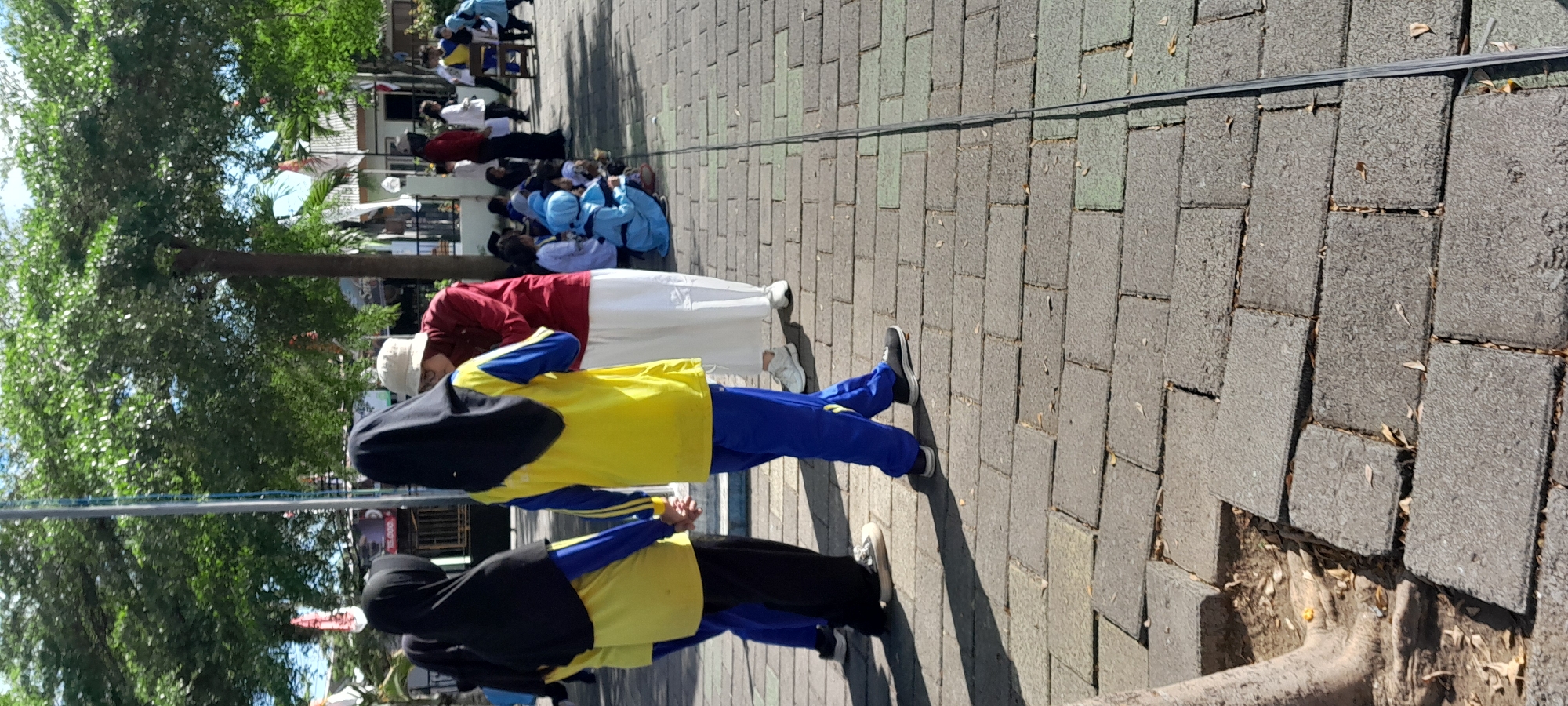 The image depicts an outdoor scene in the city of Rogojampi, Indonesia, likely on a paved walkway or plaza. In the foreground, two individuals wearing yellow and blue uniforms are holding hands. They are facing away from the camera, with one wearing a black hijab and the other partially visible. To the right, another person in a maroon top and white skirt is interacting with a group of children sitting on the ground. The children are dressed in blue and white attire, some wearing hijabs. Further in the background, other individuals are standing and walking, and a tree with lush green foliage is prominent. The ground is covered with interlocking rectangular paving stones. The environment suggests a public space, possibly a school or community gathering area, with buildings and flags visible in the distance under a bright sky.