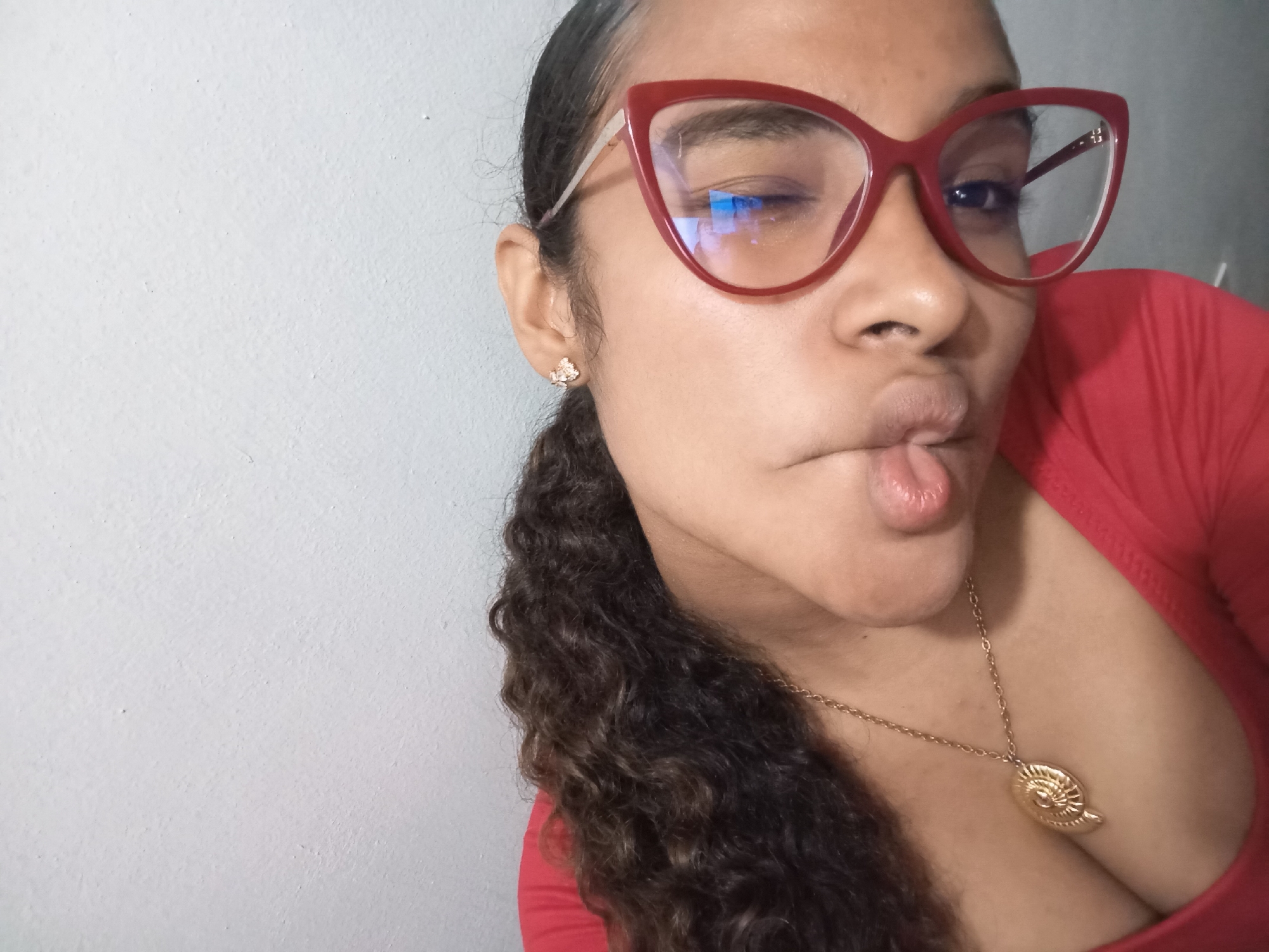 A woman is posing for a selfie. She is wearing red glasses and a red top. She has curly dark hair pulled back on one side. She is sticking her tongue out slightly and winking one eye. She is wearing a gold necklace with a large pendant. She is inside, standing in front of a plain white wall. The location is not discernible from the image.
